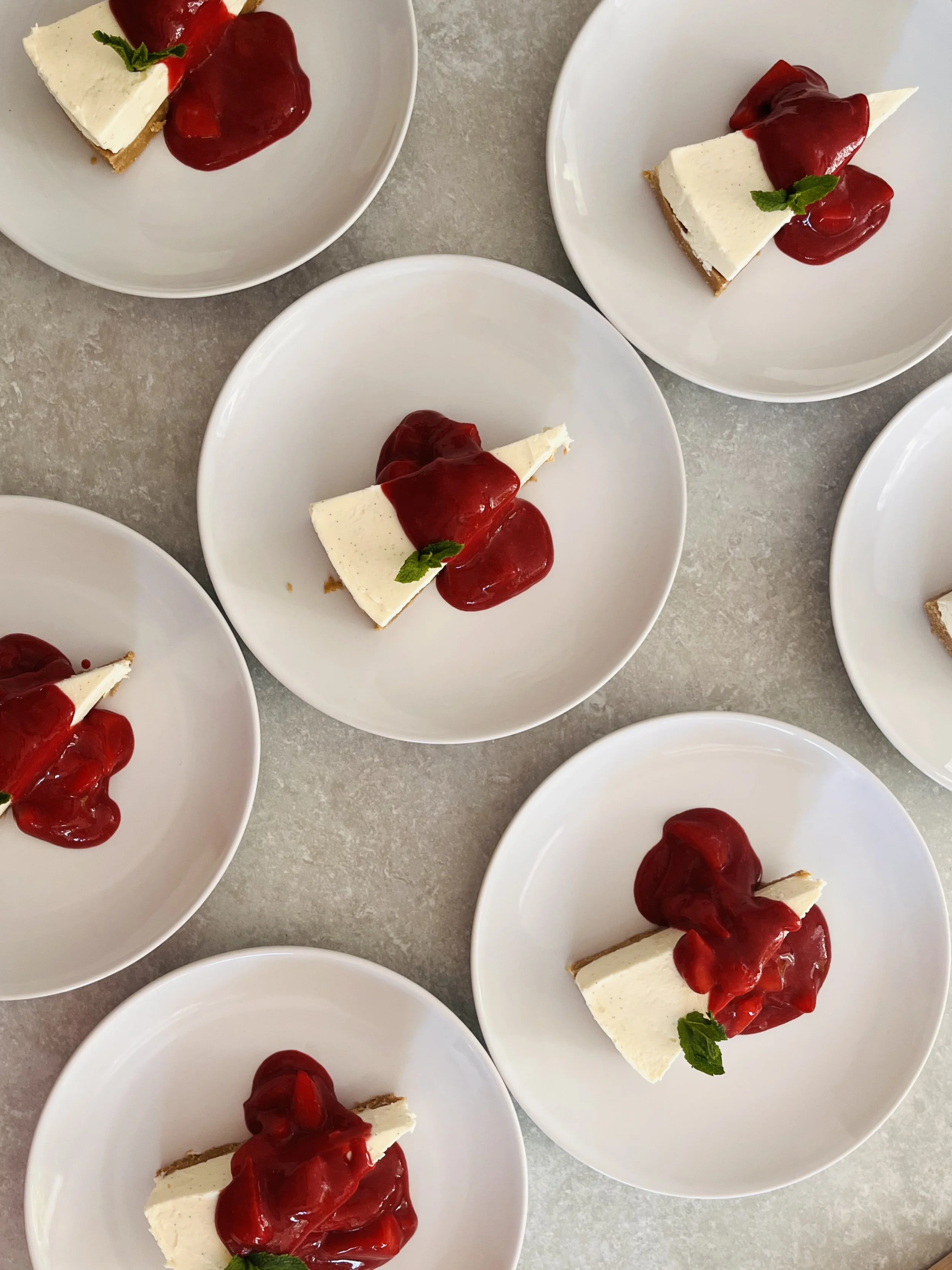 Multiple white plates each holding a slice of cheesecake topped with red cherry sauce and a small green mint leaf.