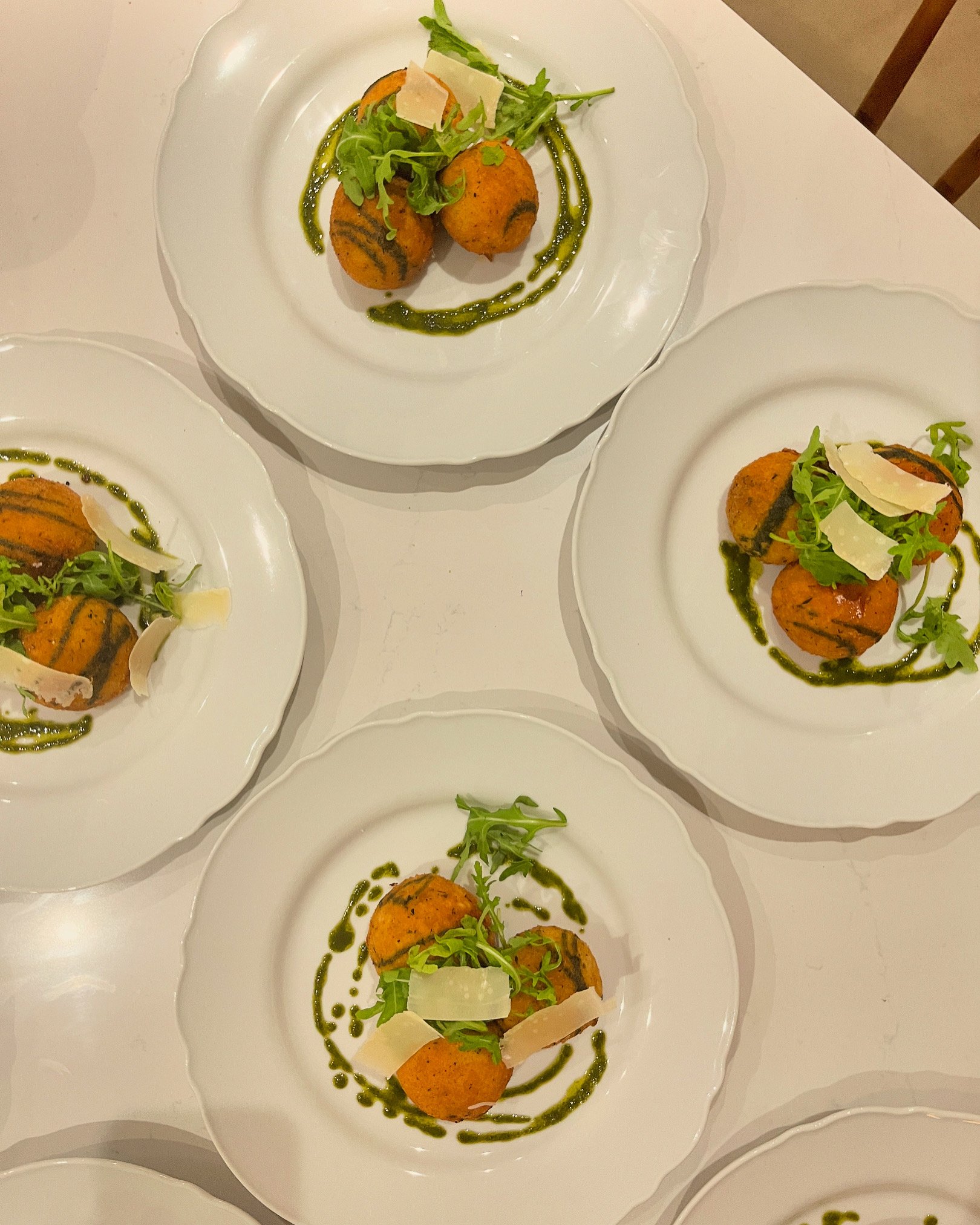 A white plate with three fried balls topped with arugula, shaved Parmesan cheese, and drizzled with green sauce, arranged in a line on a white table.