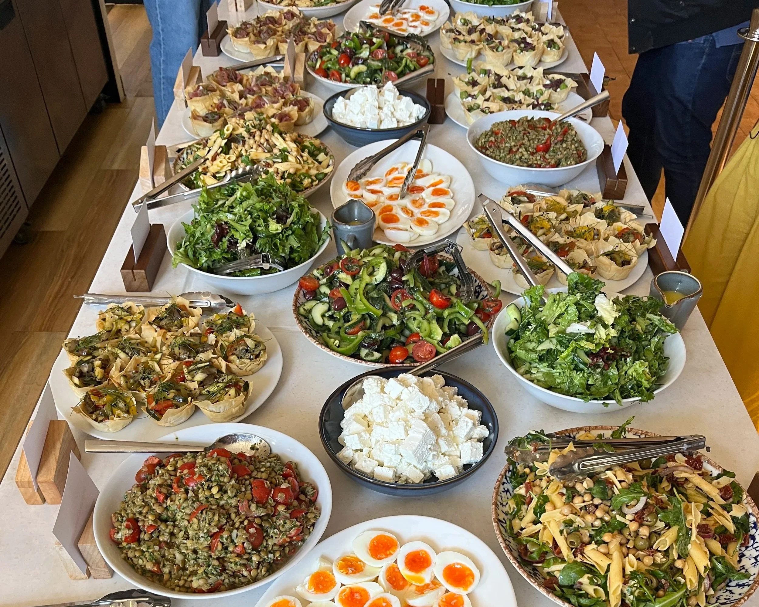 A buffet table with a variety of salads, including pasta salad, fruit salad, mixed greens, and chopped vegetables, along with bowls of crumbled cheese and halved boiled eggs.