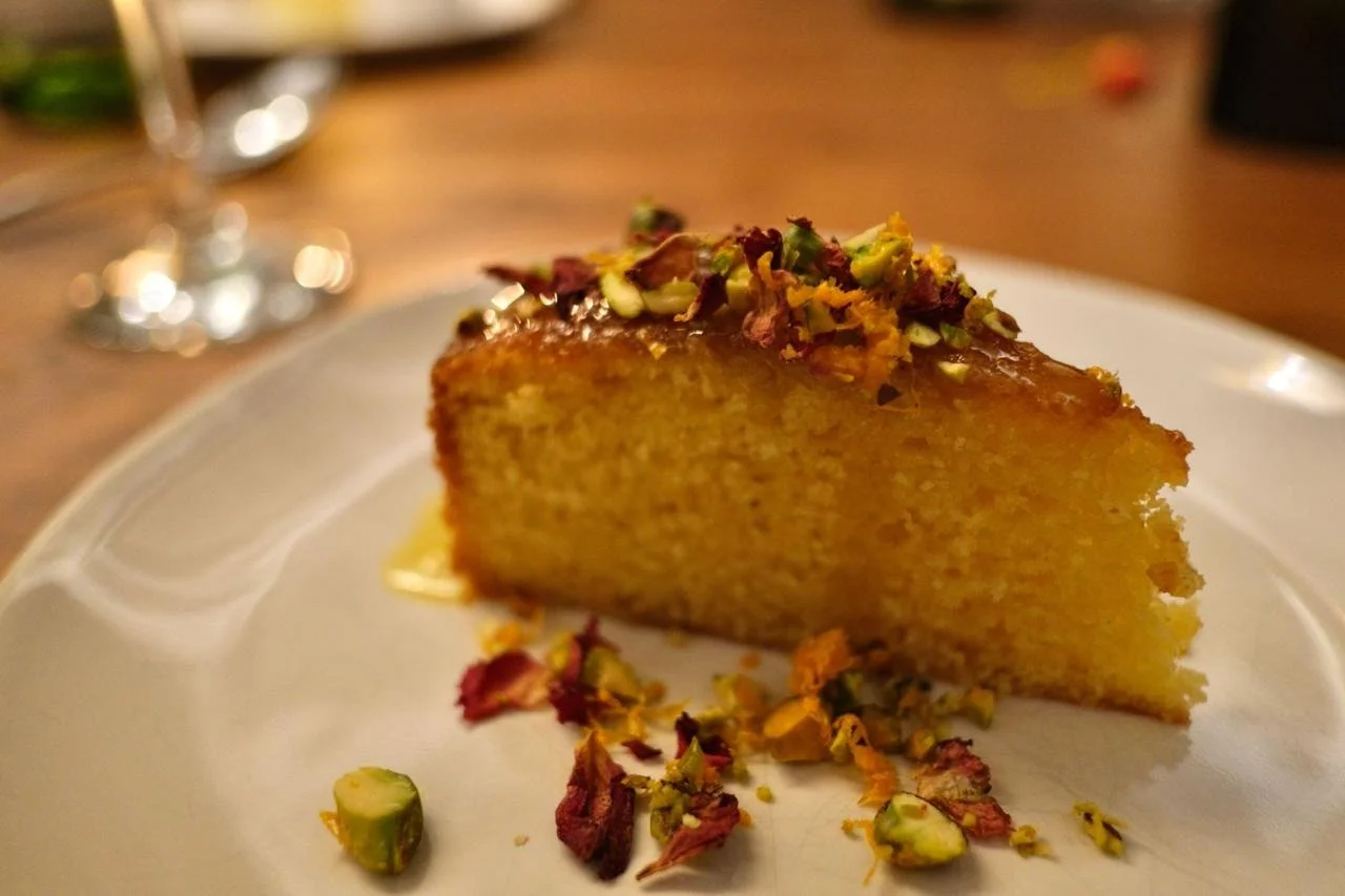 A slice of yellow cake garnished with chopped pistachios and crushed rose petals on a white plate.