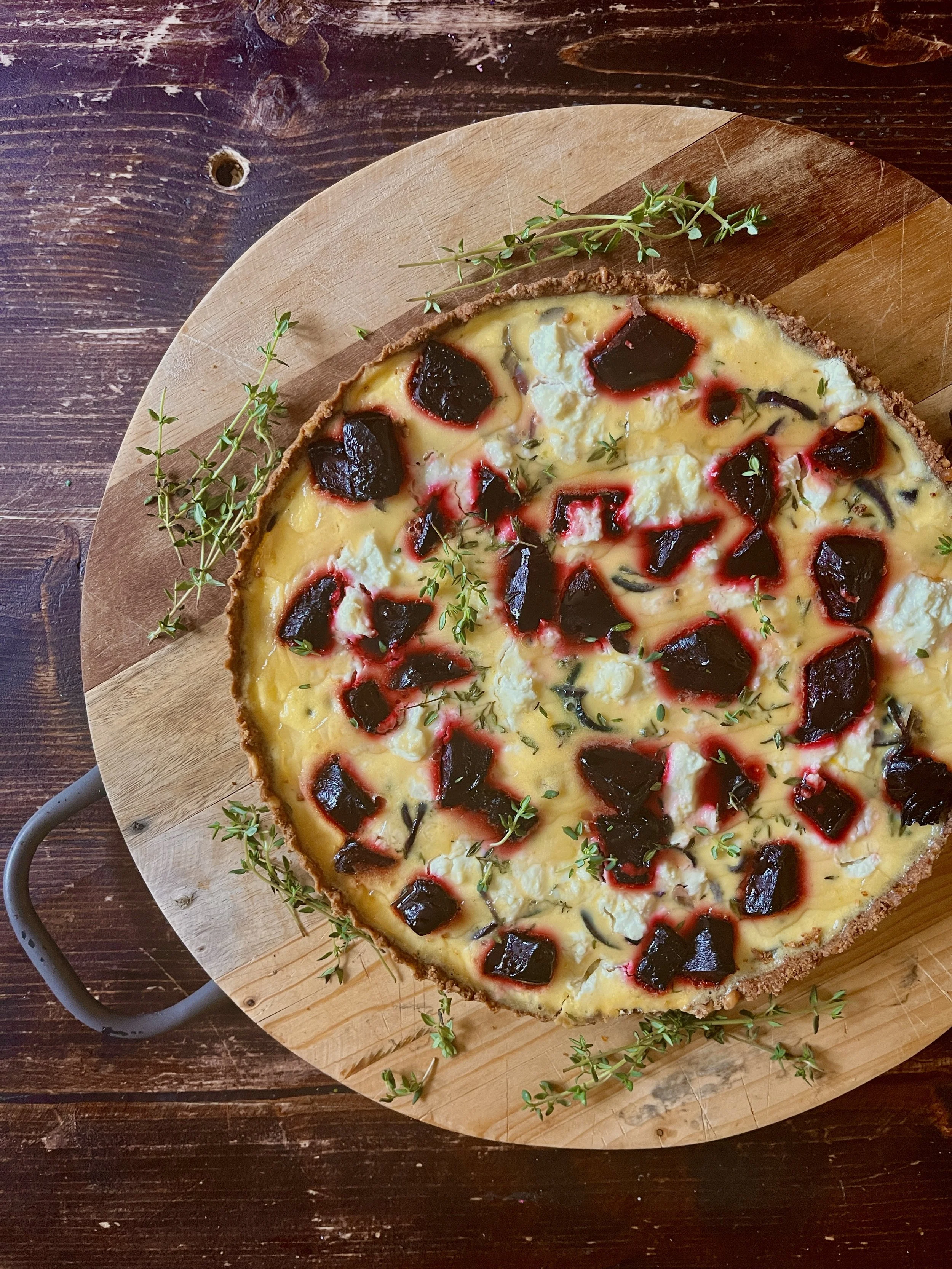 A round pizza with cheese, black olives, and small green herbs on a wooden pizza board, garnished with sprigs of fresh thyme.