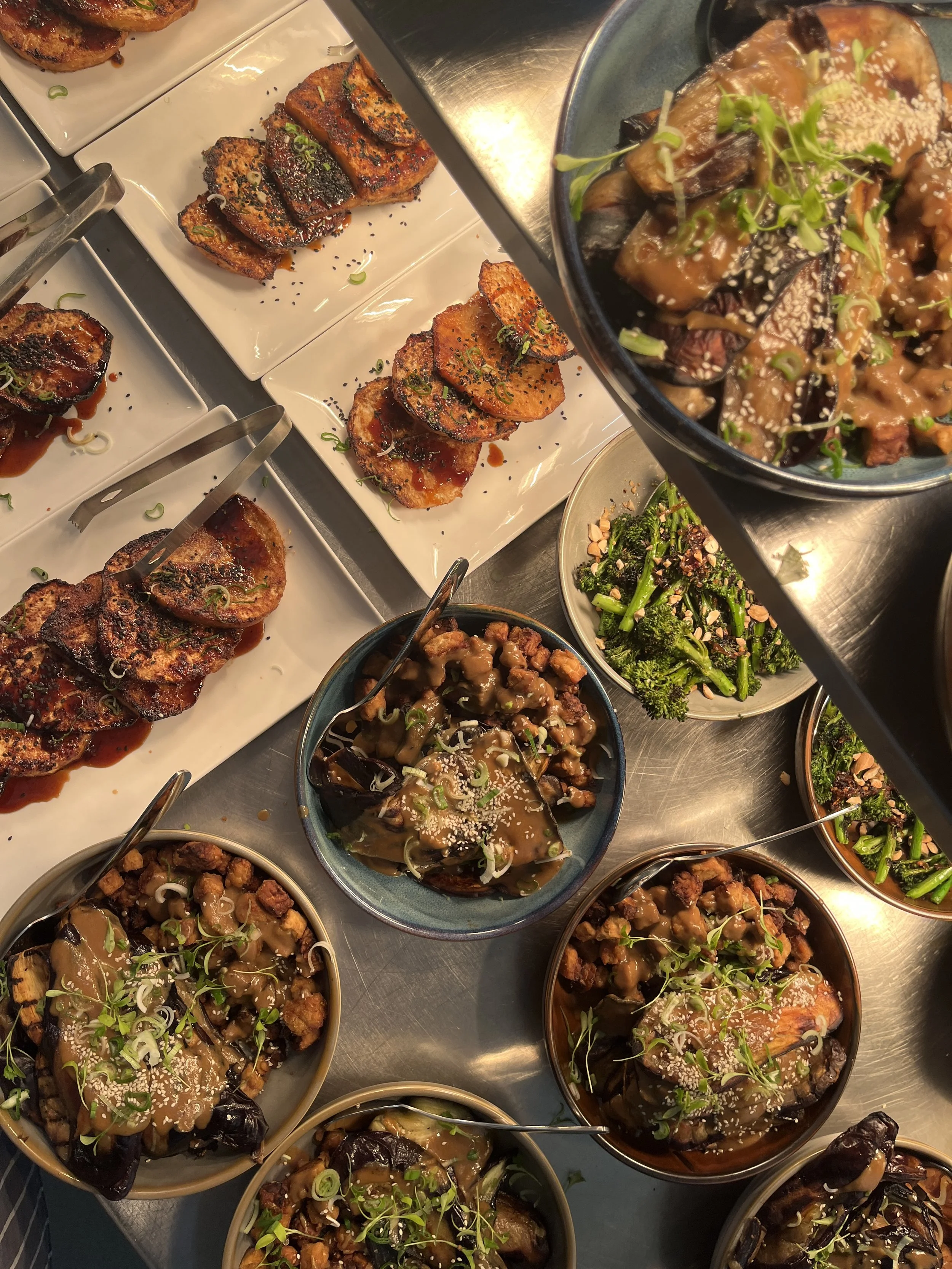 Assorted Asian dishes including eggplant, tofu, and vegetables garnished with sesame seeds and green onions, served in bowls and plates.