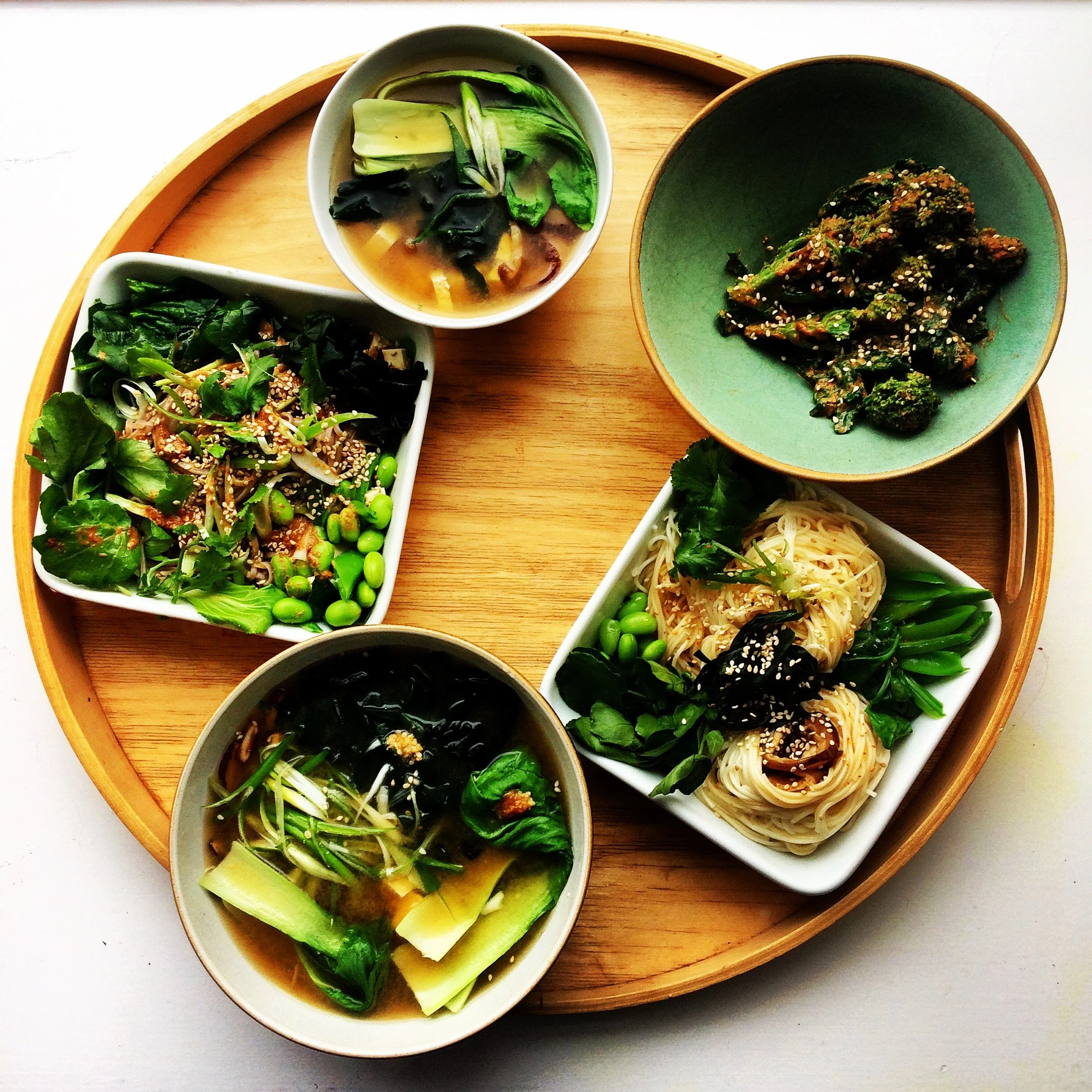 Various Asian dishes served in small bowls on a round wooden tray. The dishes include vegetables, noodles, seaweed salad, and a soup with bok choy and seaweed.