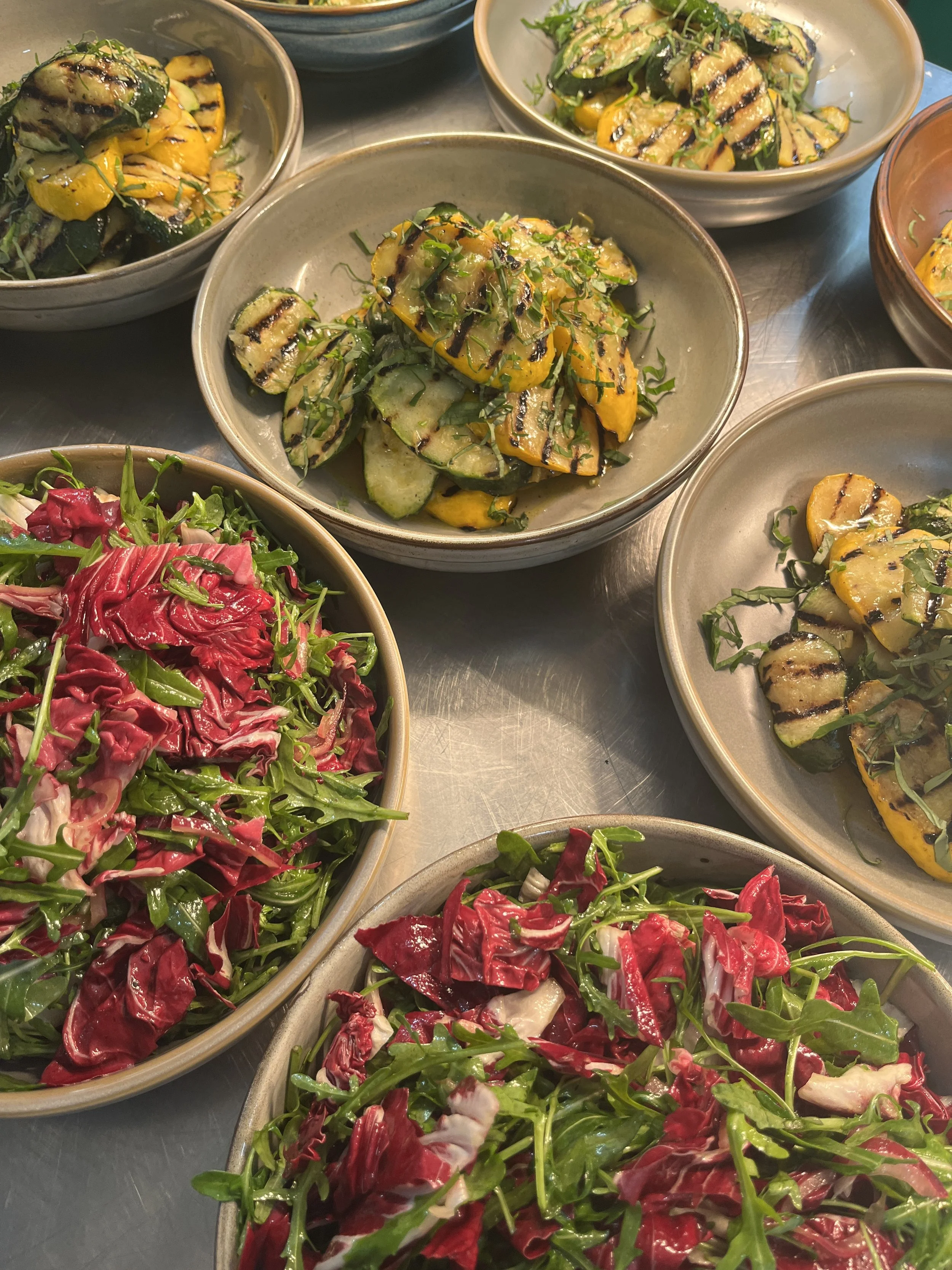 Bowls of grilled zucchini and yellow squash topped with chopped herbs, alongside bowls of mixed green and radicchio salad.