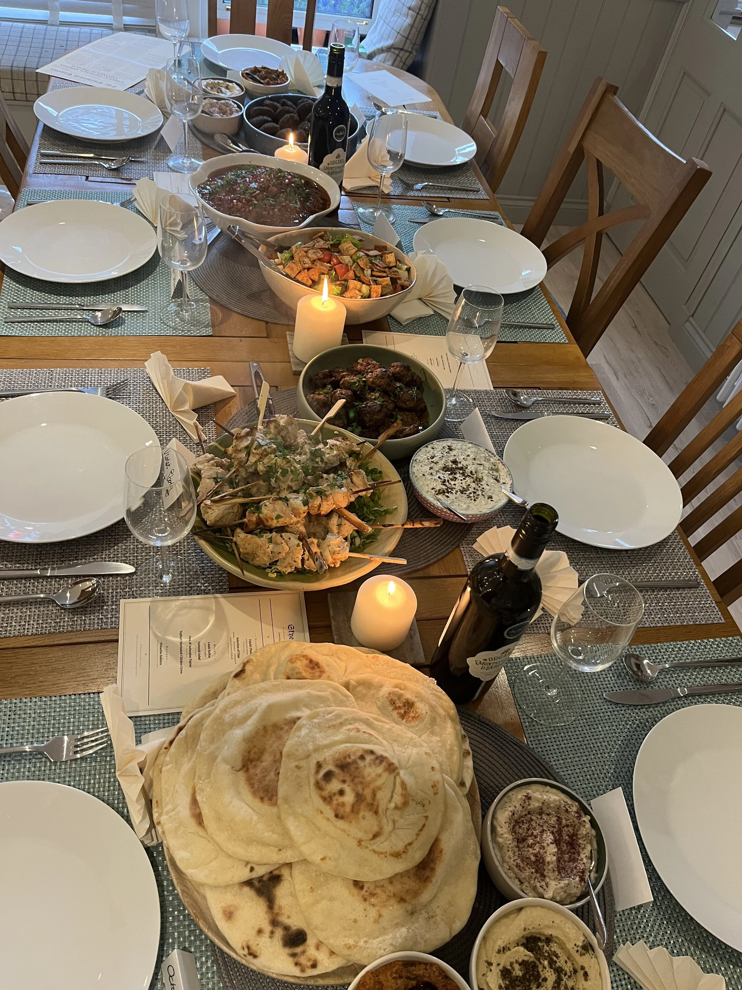 A dining table set for a meal with several dishes including naan bread, bowls of dip, a large bowl of salad, a bowl of meatballs, a bowl of stew or curry, and various utensils, glasses, and candles.