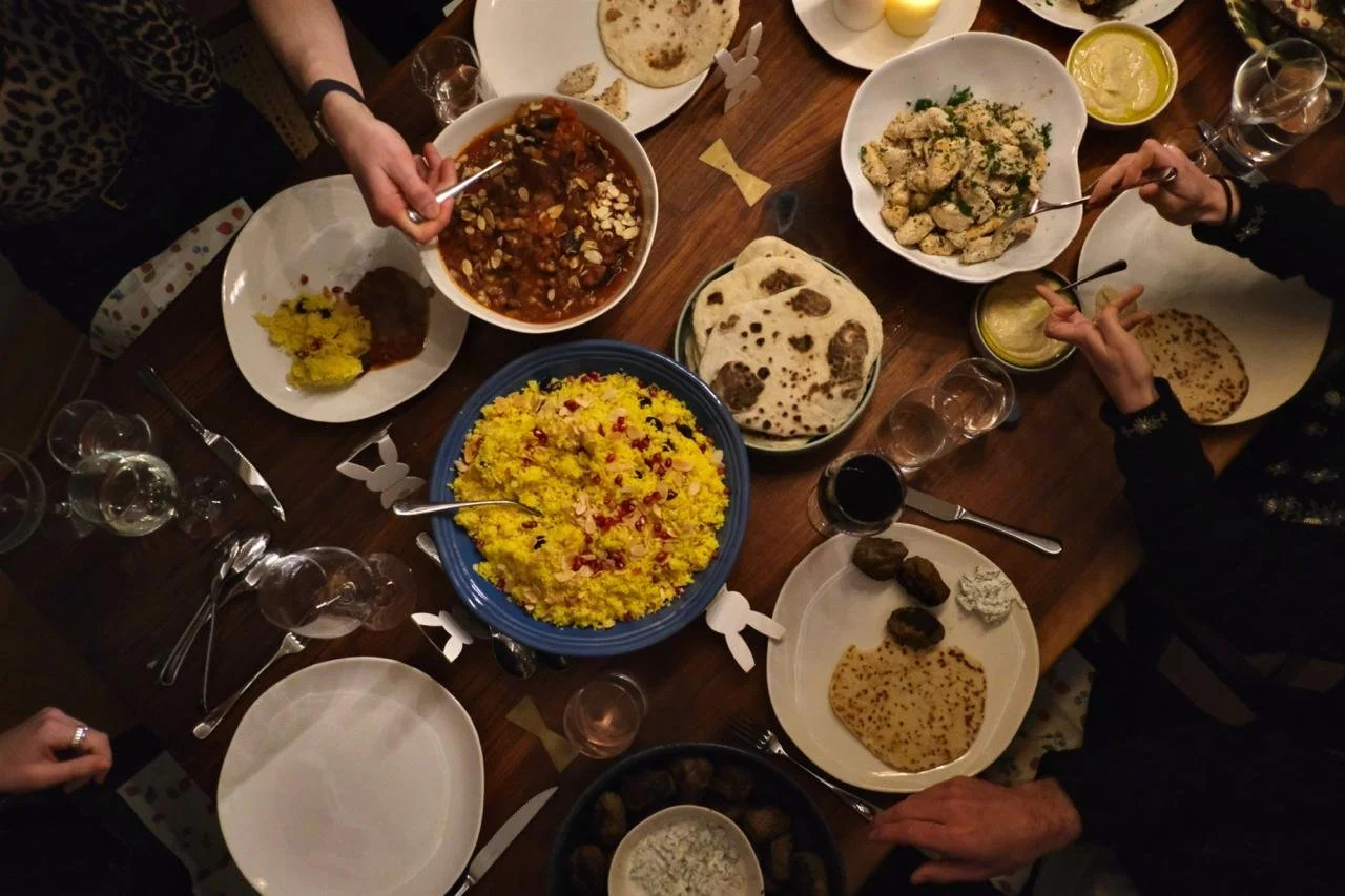 A dinner table with various Indian dishes, including naan bread, rice, curry, kachori, and drinks, with people serving and reaching for food.