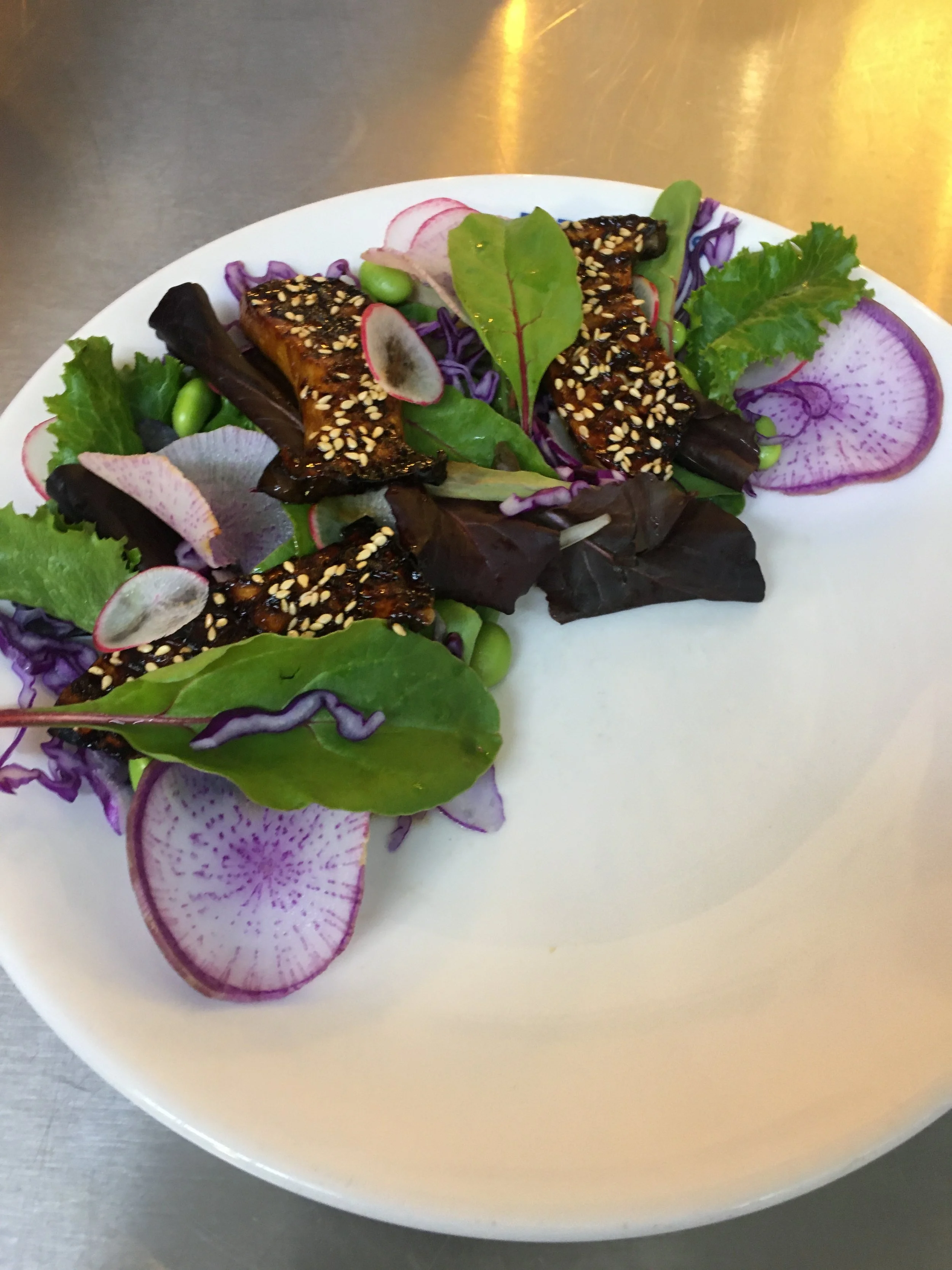 A salad with mixed greens, purple radish slices, edamame, and grilled meat with sesame seeds on a white plate.