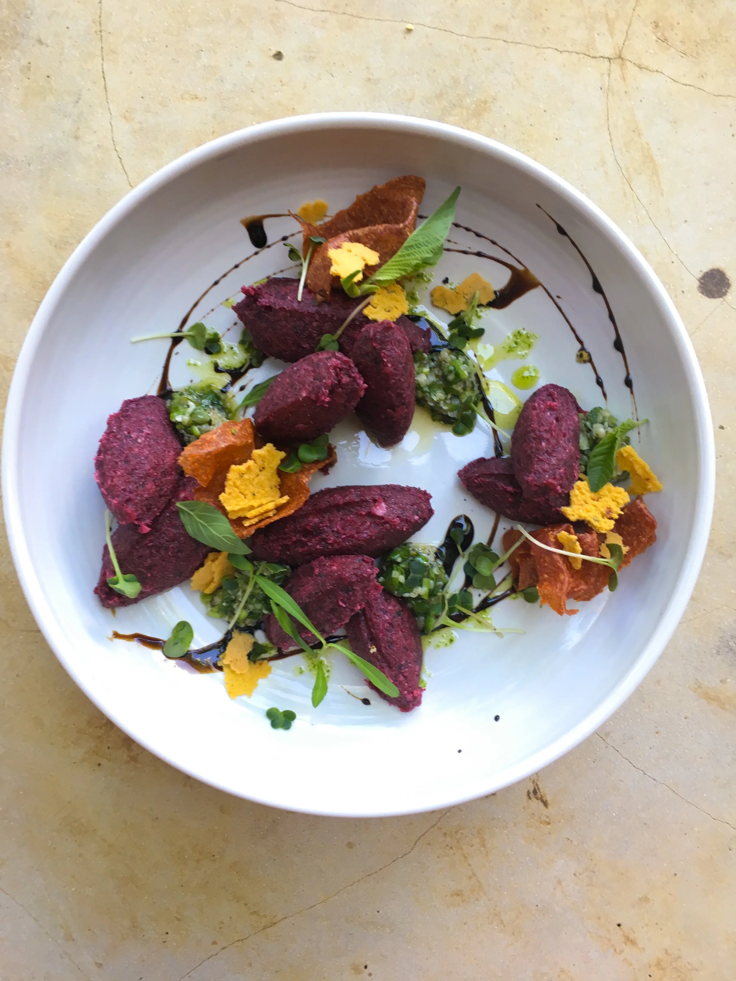 A white plate with several pieces of purple-hued food, garnished with microgreens, yellow flower petals, green herbs, and drizzled with dark and green sauces, on a beige marble surface.