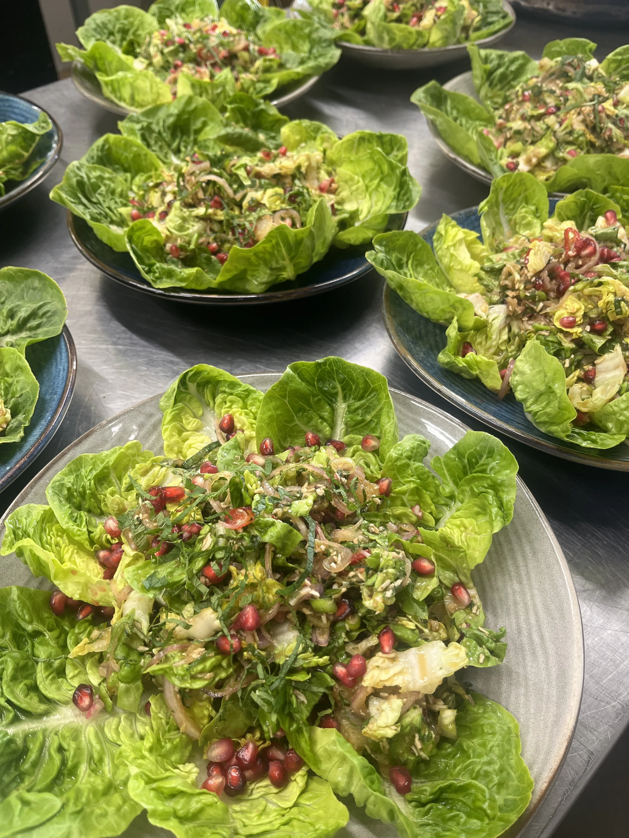Fresh green lettuce salads topped with pomegranate seeds, served in various bowls on a gray table.