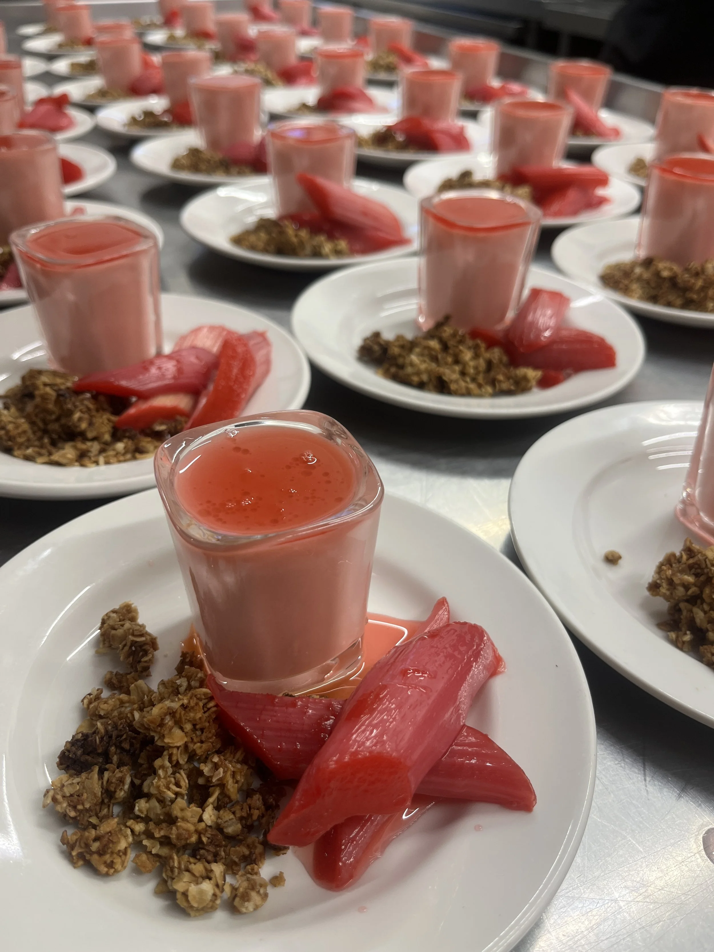 Multiple white plates with pink shot glasses filled with a pink liquid, sliced strawberries, and granola are arranged on a stainless steel counter.
