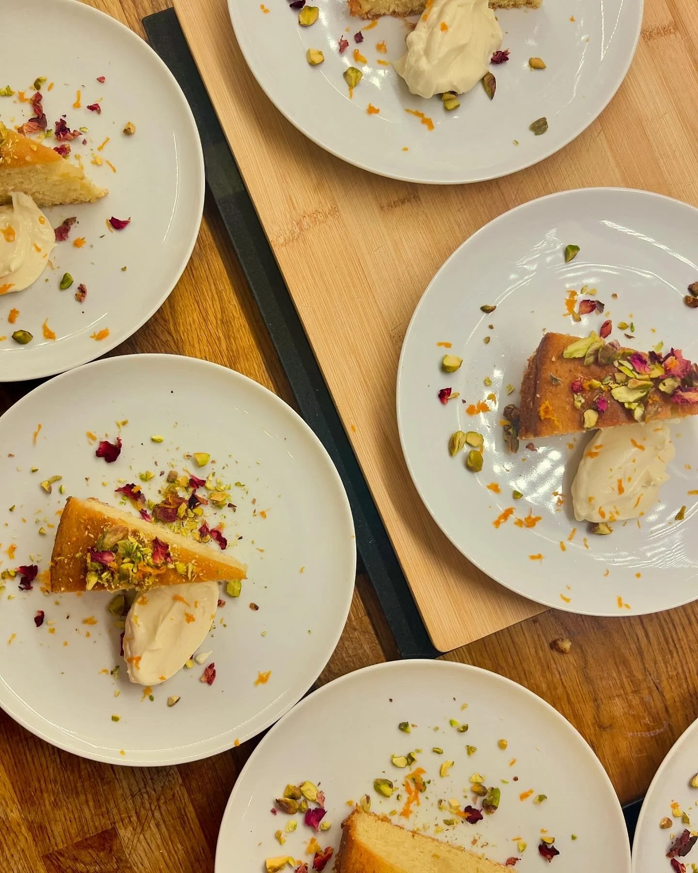 Several white plates with slices of cake, ice cream, and sprinkled pistachios and rose petals, arranged on wooden tables.