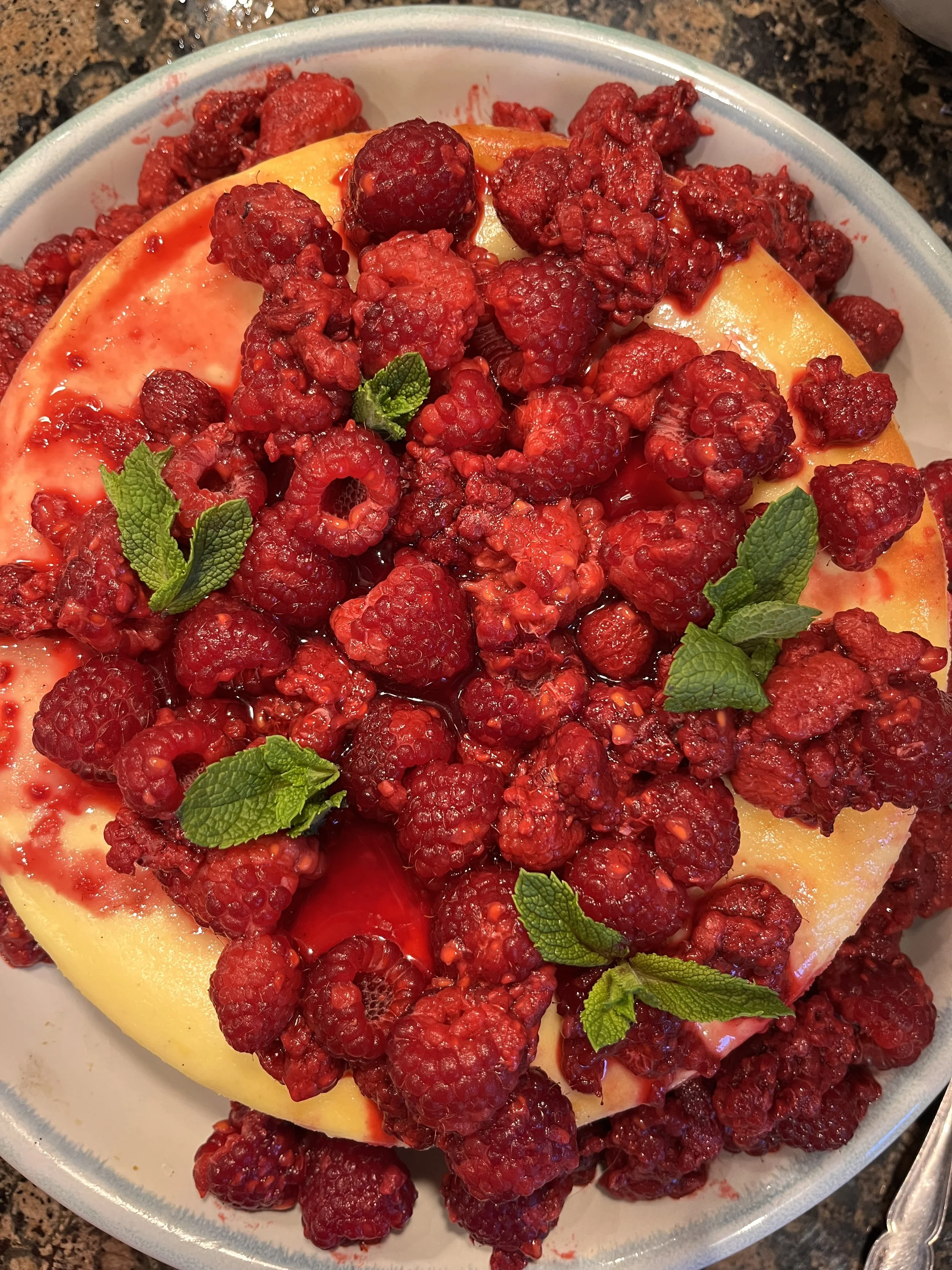 A close-up image of a dessert topped with raspberries and mint leaves, with a yellow base layer, possibly cheesecake or mousse, on a white plate.