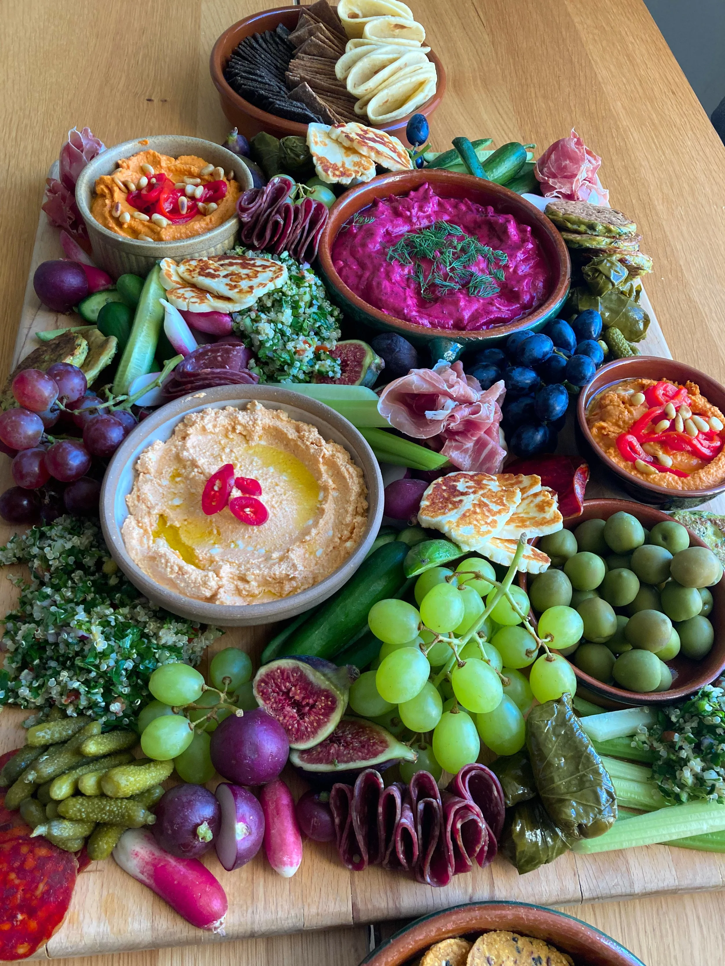 Wooden table with a colorful array of fresh fruits, vegetables, hummus, cheese, crackers, and dips arranged in bowls and on the table. Items include grapes, figs, radishes, pickles, kohlrabi, and an assortment of cheeses and spreads.