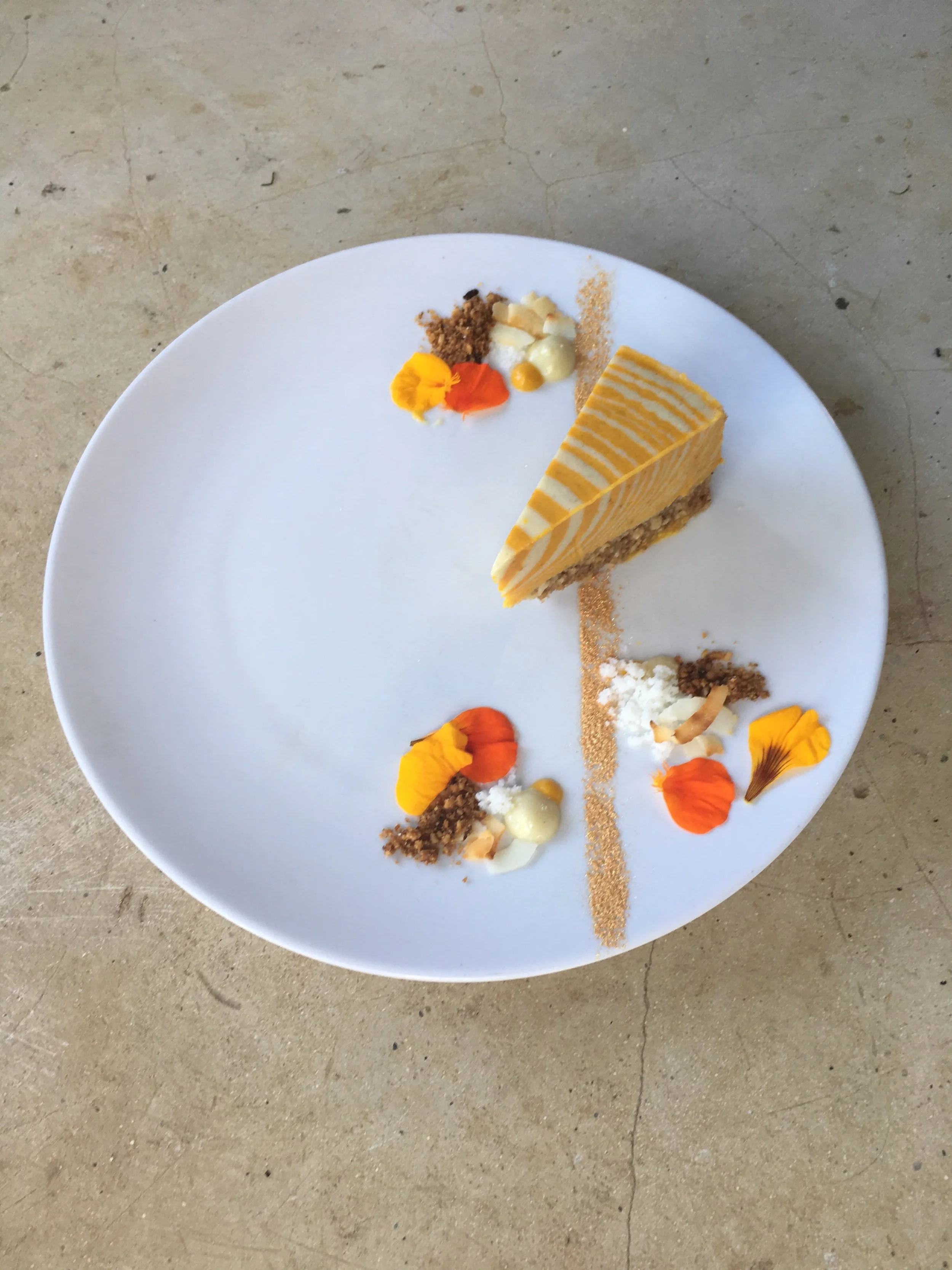 A white plate with a slice of layered yellow and orange dessert, garnished with edible flowers and small dollops of cream, with decorative gold and white accents on a concrete surface.