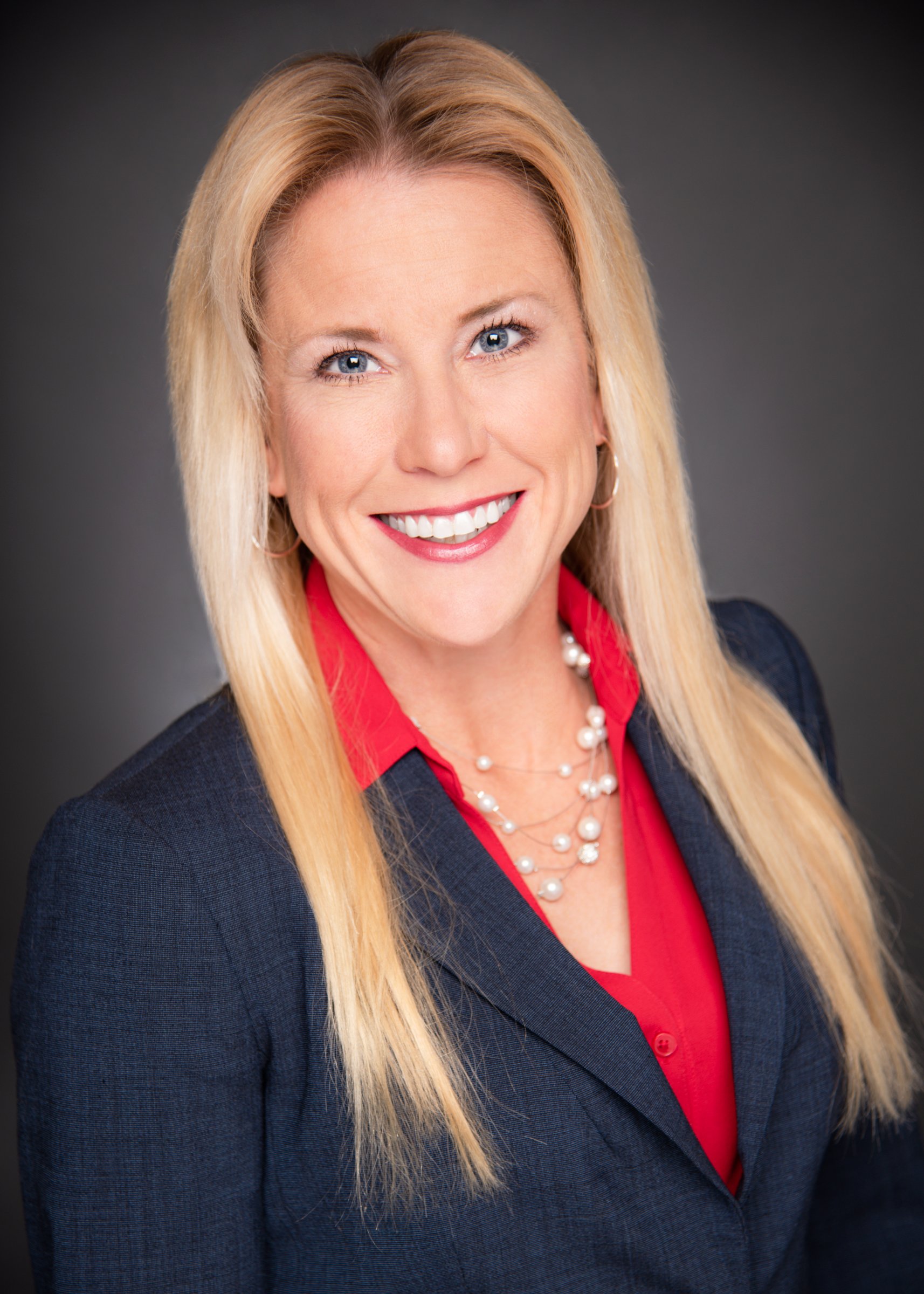 Headshot of a smiling woman with long blonde hair, wearing a navy blazer, red blouse, pearl necklace, and earrings, against a dark background.