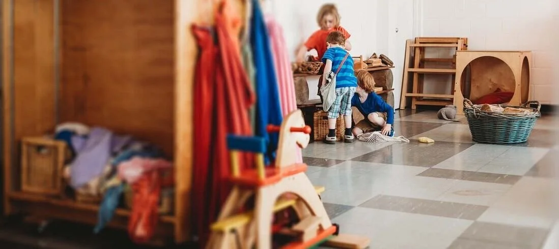 Four young children playing together in a brightly lit indoor play area with wooden furniture and toys.