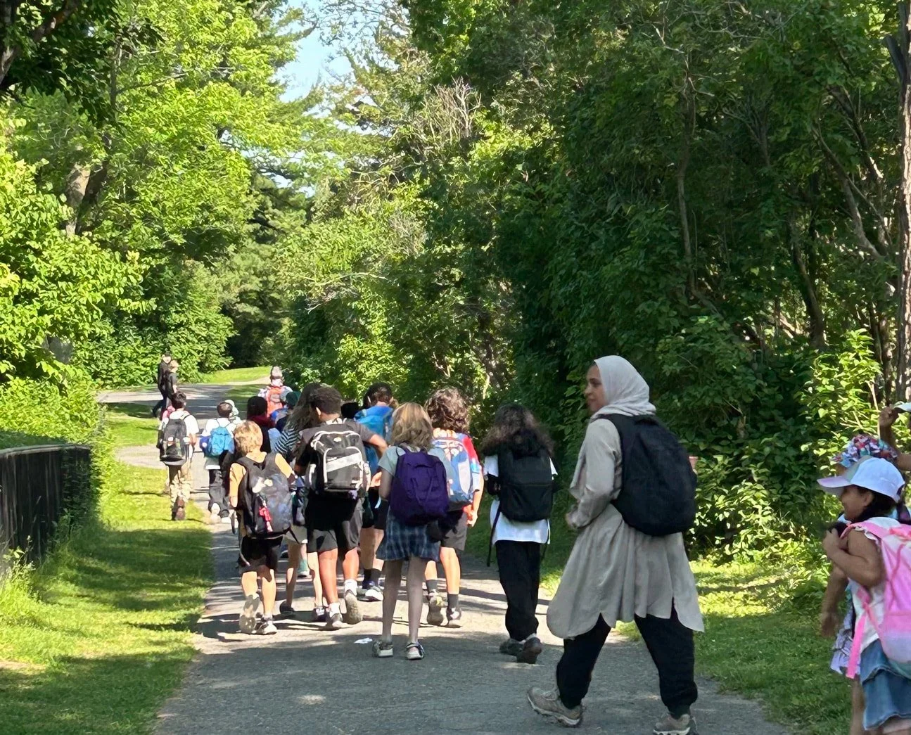 A group of children and adults walking on a park trail surrounded by green trees and bushes.
