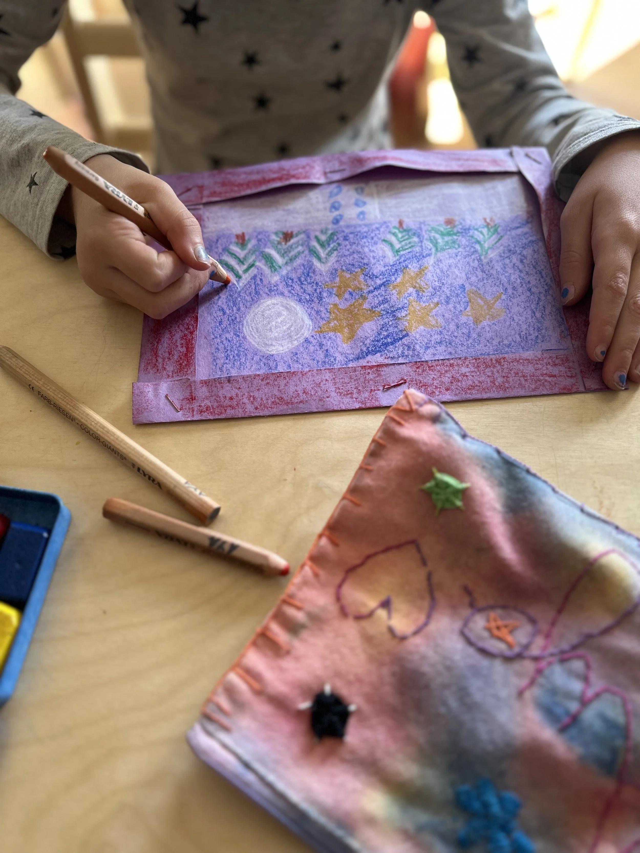 A child's hand is coloring on a pink paper with some materials around including colored pencils, fabric pieces, and a coloring set. The paper features stars, a moon, and decorative patterns.