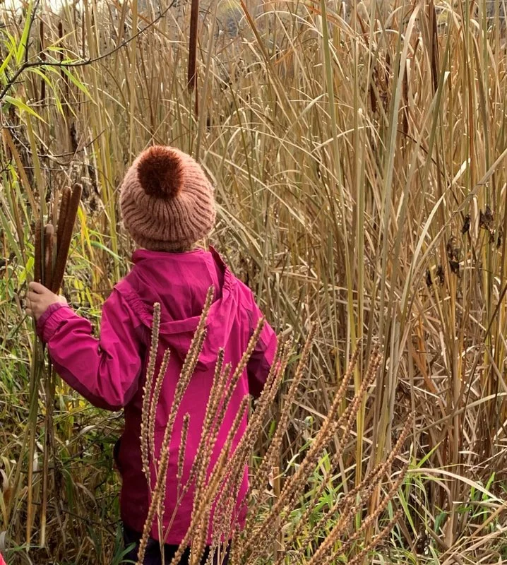 Child in pink jacket and knit hat exploring a tall grassy field, holding sticks.
