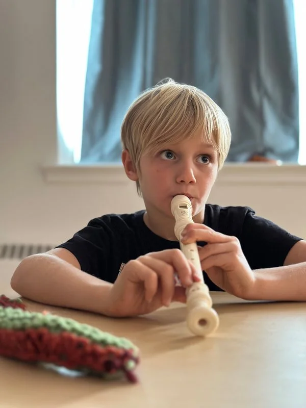 A young boy with blond hair wearing a black shirt playing a recorder at a table indoors.