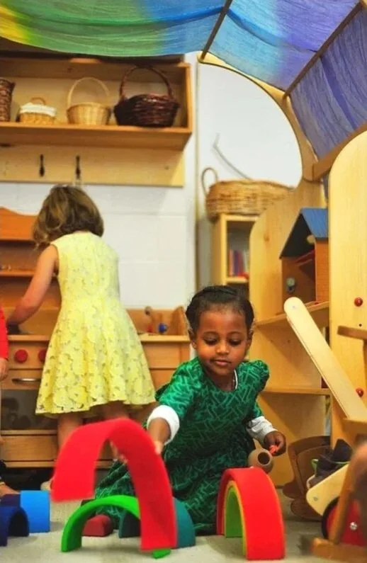 Children playing in an indoor play area with wooden toys, shelves, and colorful fabric ceiling.