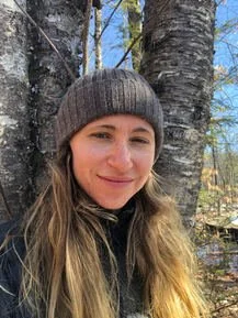 A woman with long blonde hair wearing a gray beanie and a black jacket, standing outdoors near a tree in a wooded area on a sunny day.