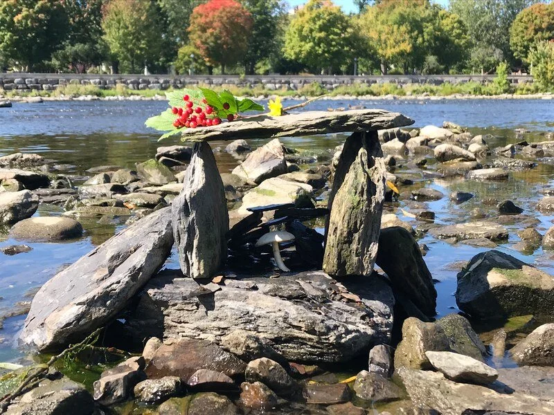 Rocks and stones build a small shelter on the riverbank, with a leaf and berries placed on top, and trees with green and orange leaves in the background.