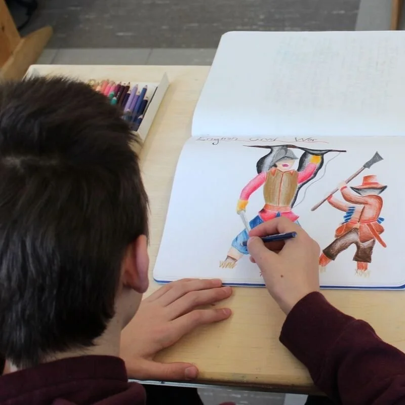 Child drawing a colorful picture of two characters in a sketchbook on a light-colored table with colored markers beside.