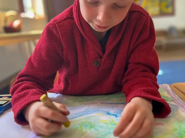A young boy in a red sweater drawing with colored markers on a sheet of paper.