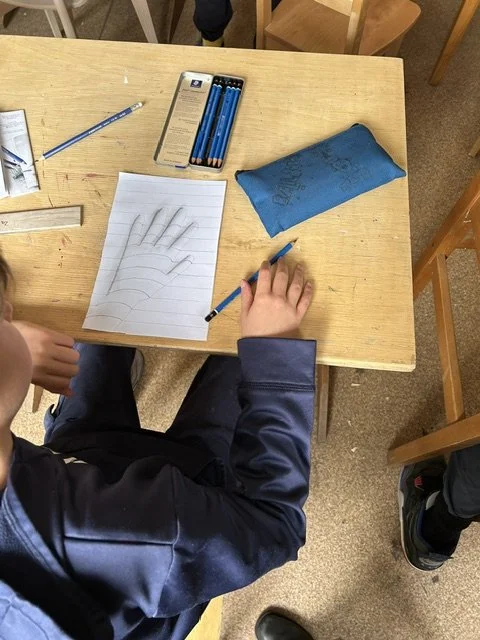 Child sitting at a desk drawing a hand on a piece of paper with a pencil, surrounded by school supplies including a blue pencil case, a set of colored pencils, and other stationery.