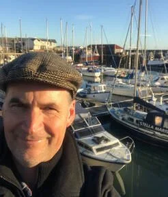 A man in a brown flat cap near a marina with several sailboats and yachts in the background.
