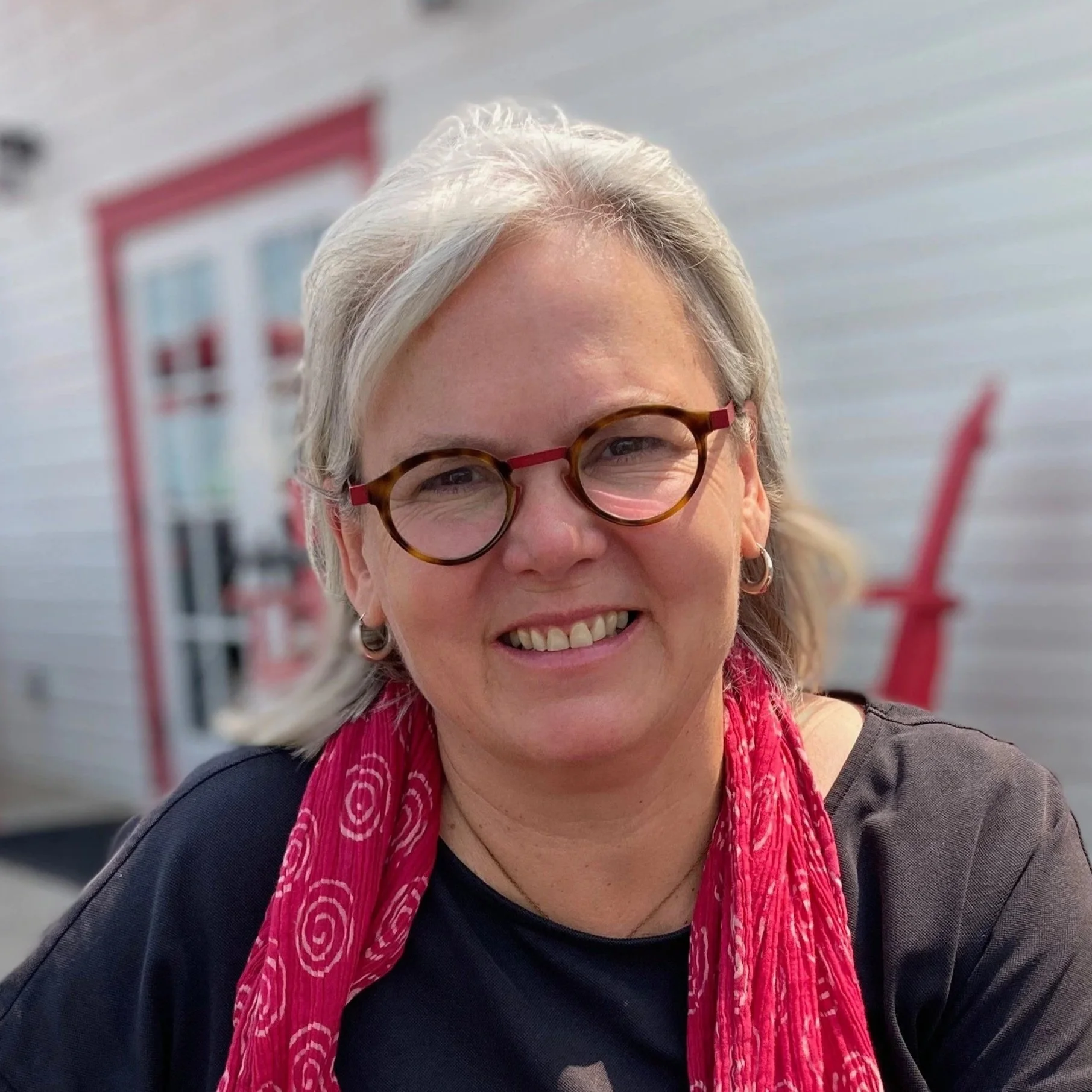 Close-up of a smiling woman with short gray hair and glasses, wearing a black top and a red patterned scarf, outdoors with a blurred background of a building and a sign.