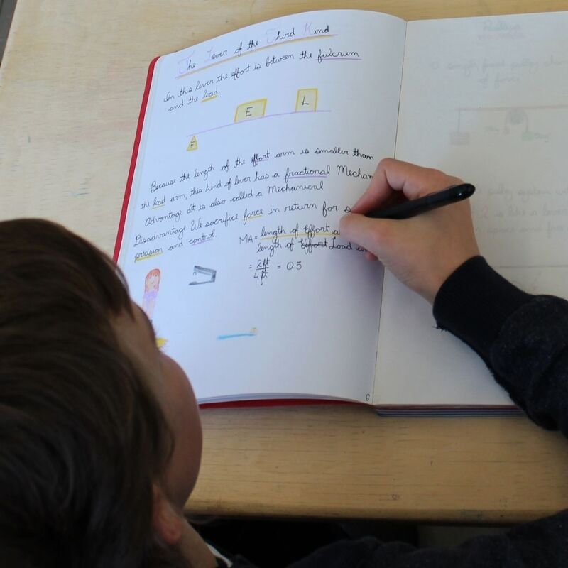 A person writing in a notebook with a black pen, with a child observing the page. The notebook contains handwritten notes and colored diagrams related to the third law of motion, including labels, formulas, and annotations.
