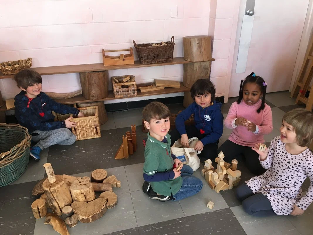 Children playing with wooden blocks and toys on the floor in a classroom or playroom.