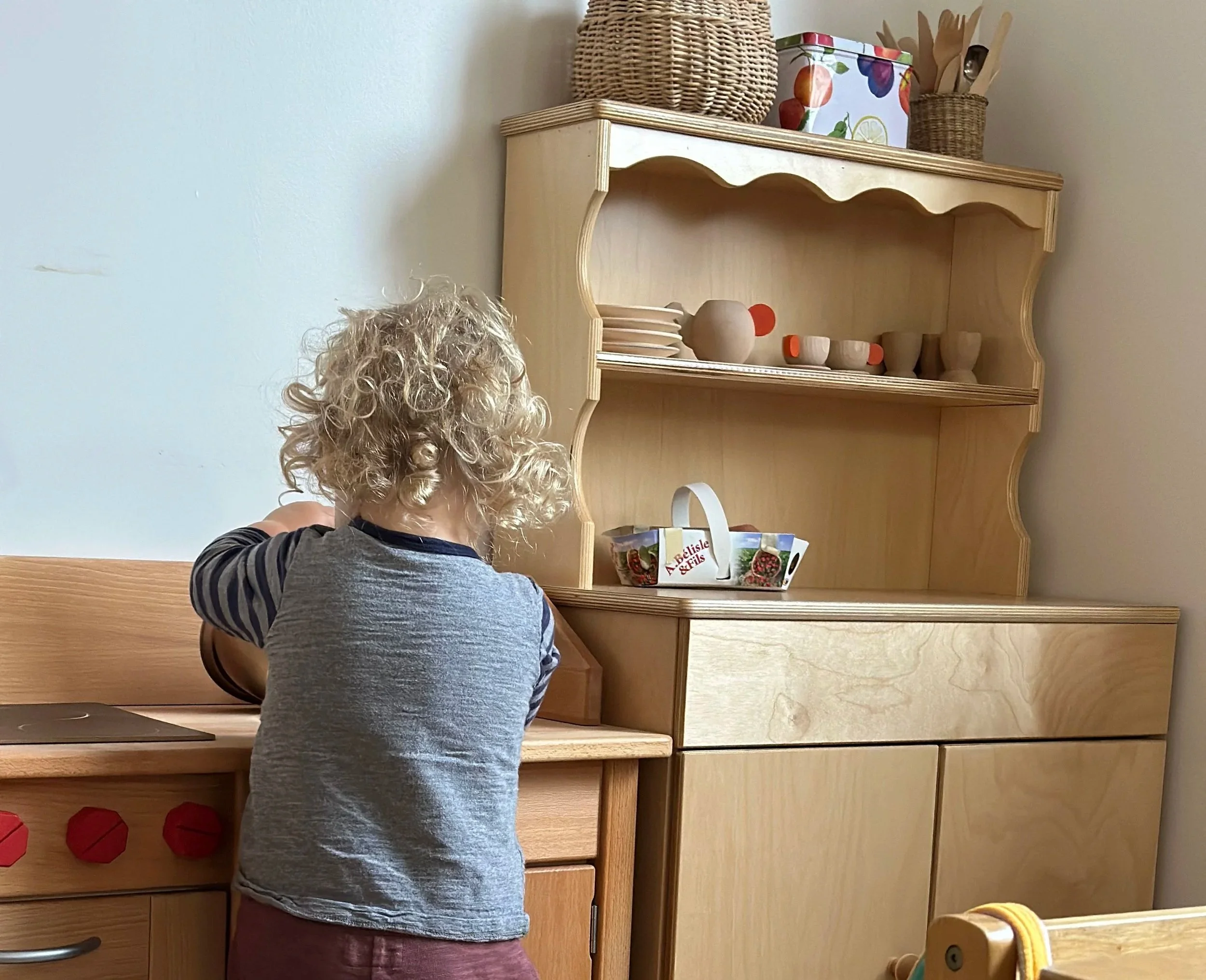 A toddler with curly blonde hair facing a light wood shelving unit that holds various items including wooden cups, a vase, a basket, and a container with a fruit pattern, in a room with light-colored walls.