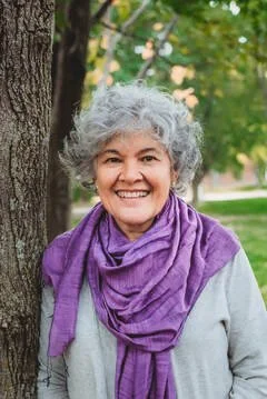 A woman with curly gray hair smiling outdoors, wearing a purple scarf and gray sweater, standing next to a tree with green foliage in the background.