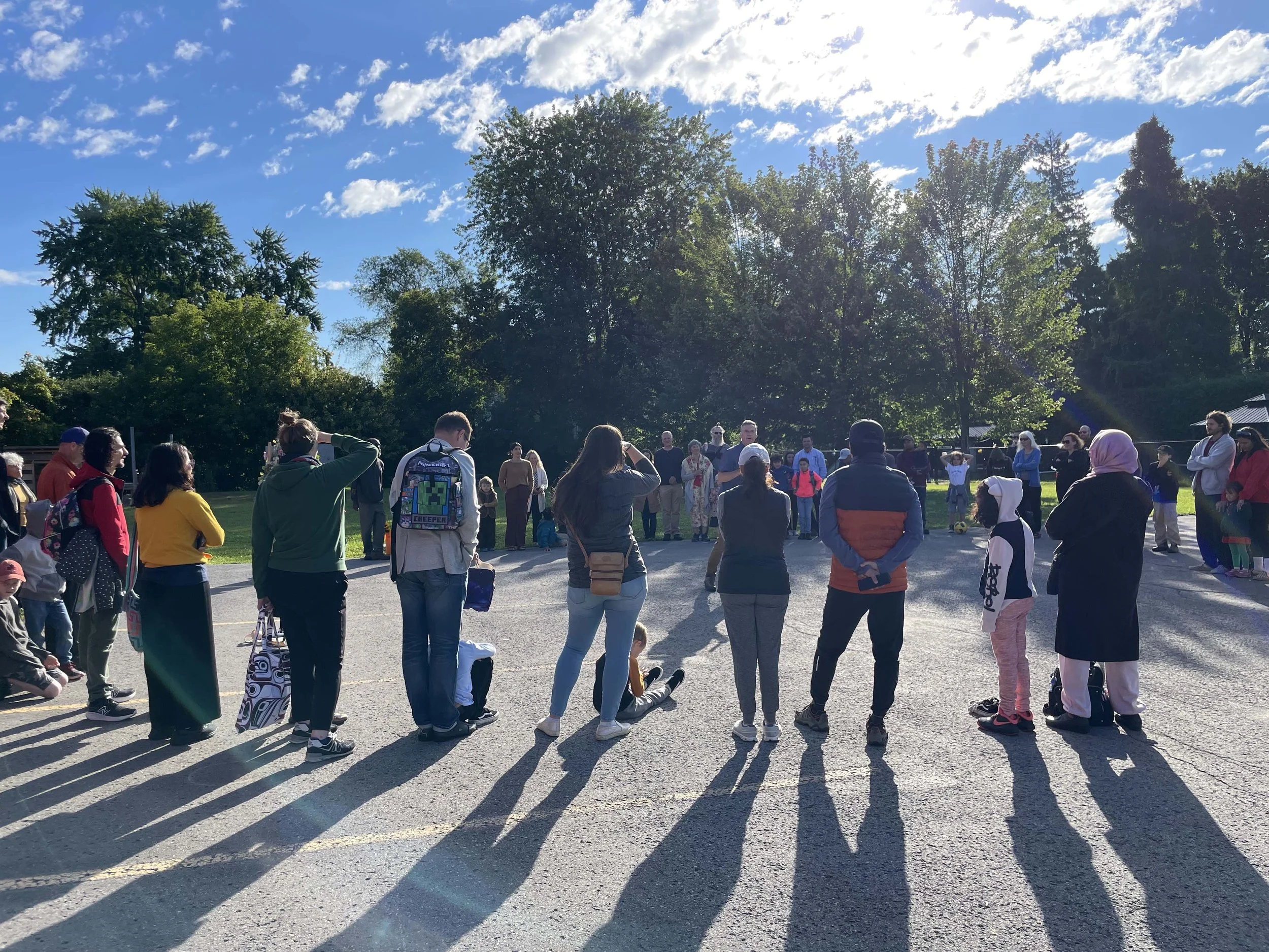 People gathered outdoors on a sunny day, standing and sitting on the ground, with large trees and a bright blue sky with scattered clouds in the background.