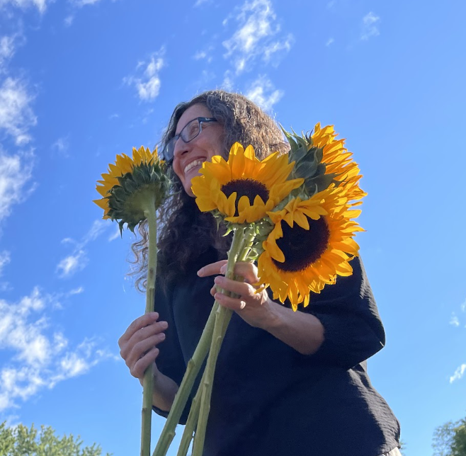 Woman with curly hair and glasses holding a bunch of sunflowers outdoors on a sunny day with a blue sky and scattered clouds.