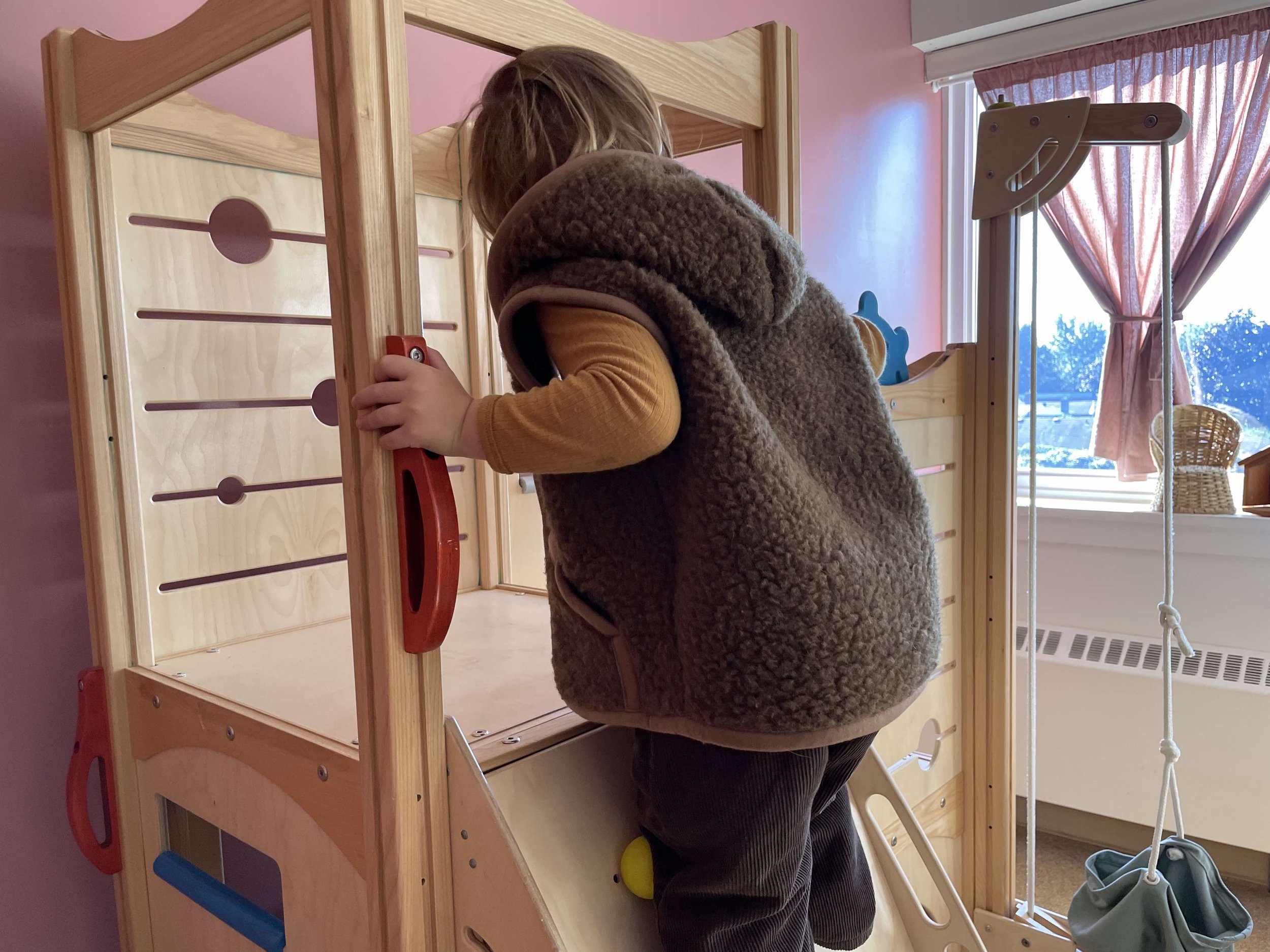 A child in a brown fuzzy vest and yellow long sleeve shirt climbs an indoor wooden play structure with a slide, in a room with pink walls and a window with partially drawn pink curtains, with trees visible outside.