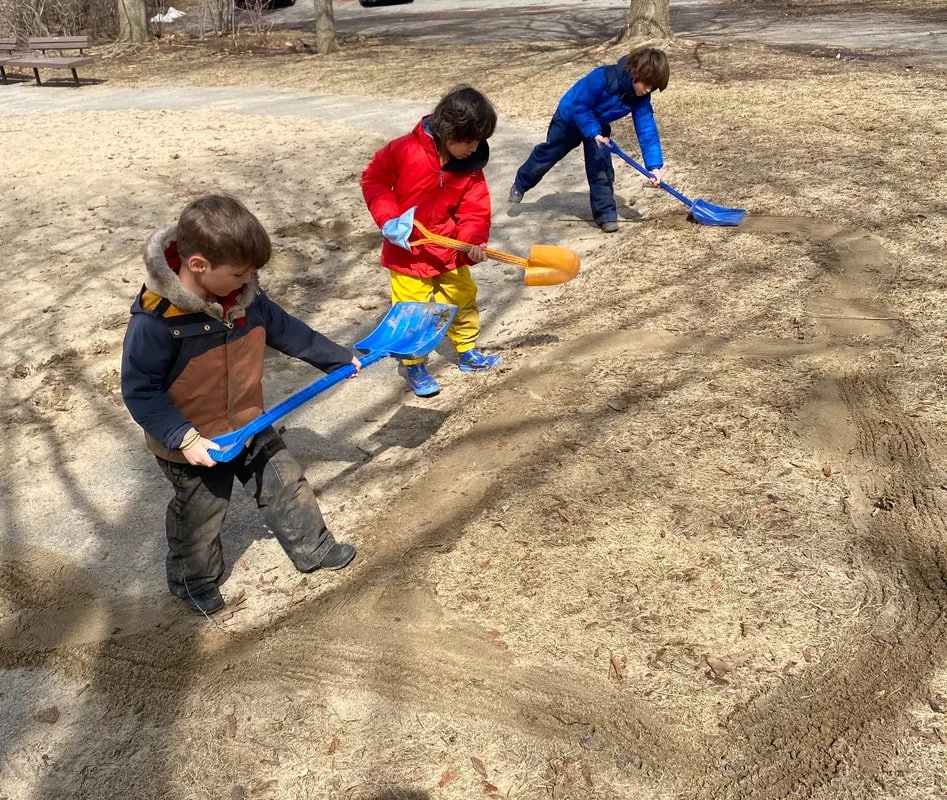 Three children playing and digging in the sand with small shovels in a park on a sunny day.