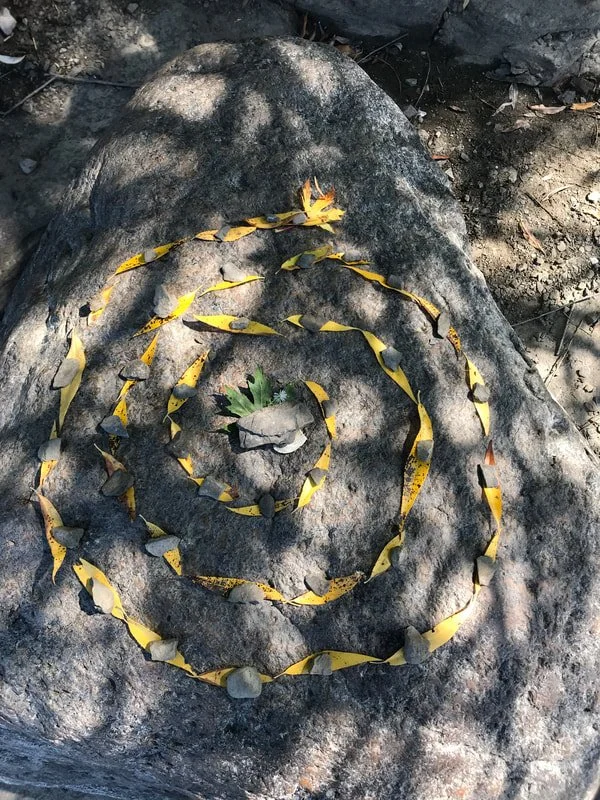 Rock with a spiral pattern made of yellow leaves and small stones, outdoors with dirt and shadow.