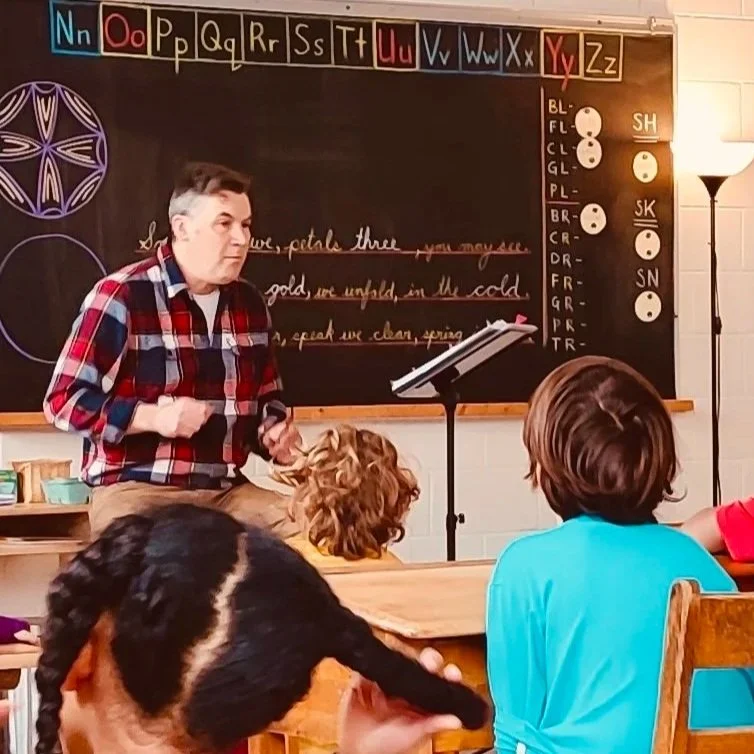 A classroom scene with a male teacher standing in front of a blackboard, teaching children. The blackboard displays the alphabet and some notes.