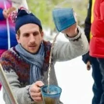 A man wearing a knit hat and scarf holds a spoon out of a bowl, with snow and people in winter clothing in the background.