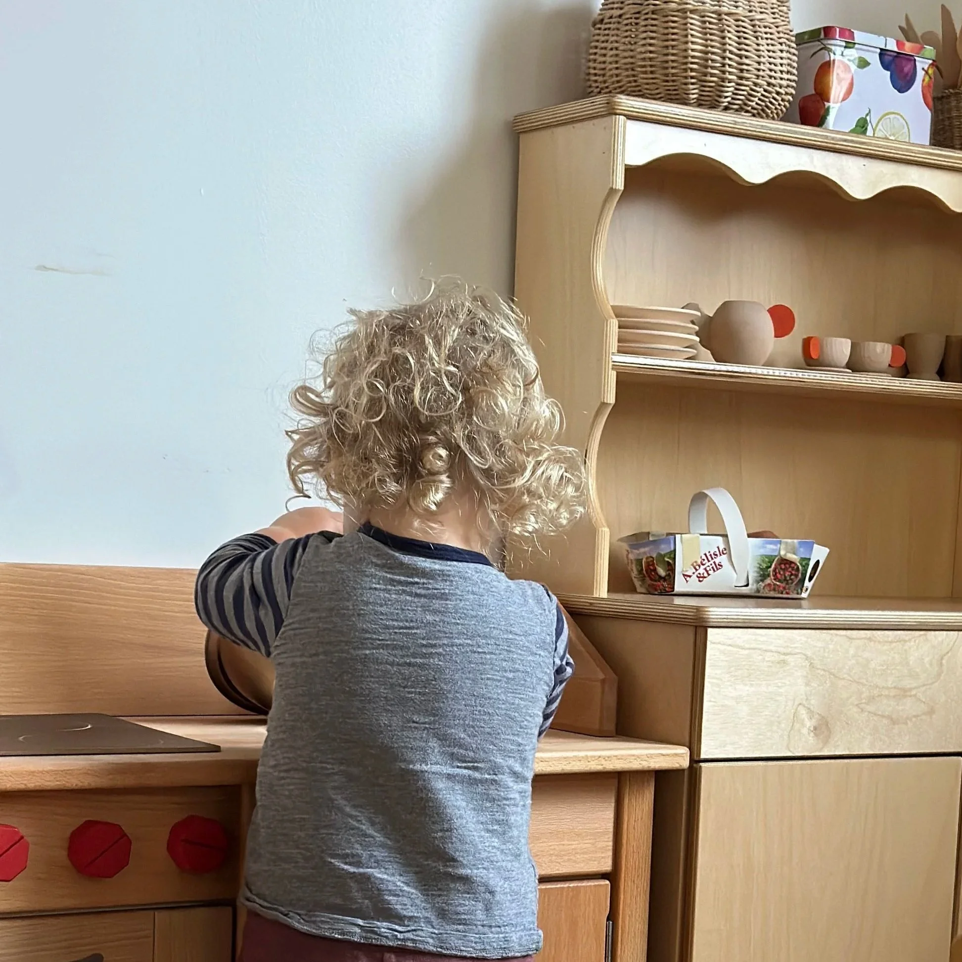 A toddler with curly hair and striped shirt standing in front of wooden kitchen with decorative items and books.