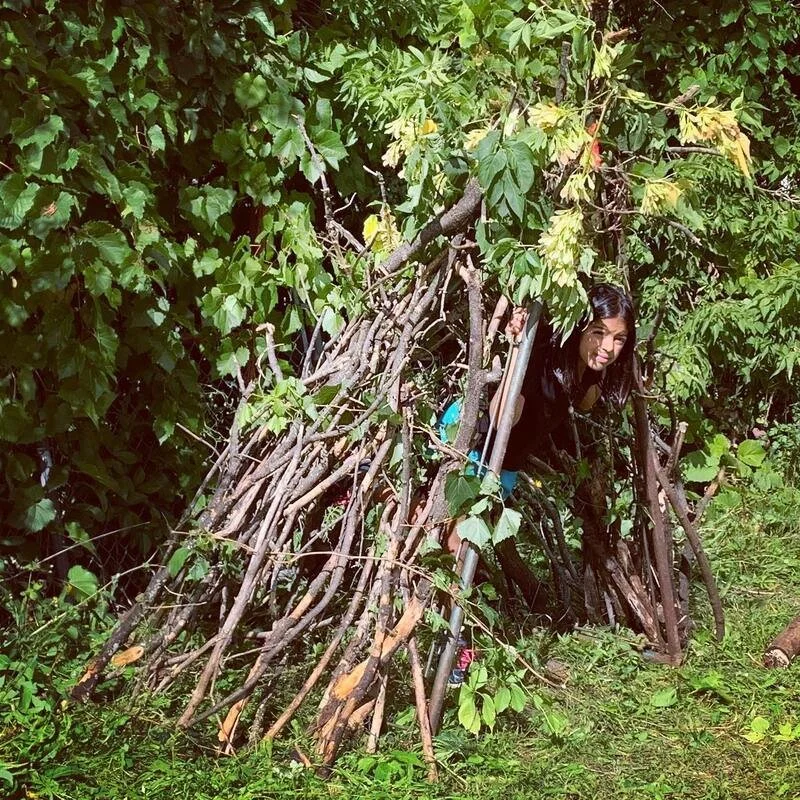 Person peeking out from a small shelter made of branches and leaves.