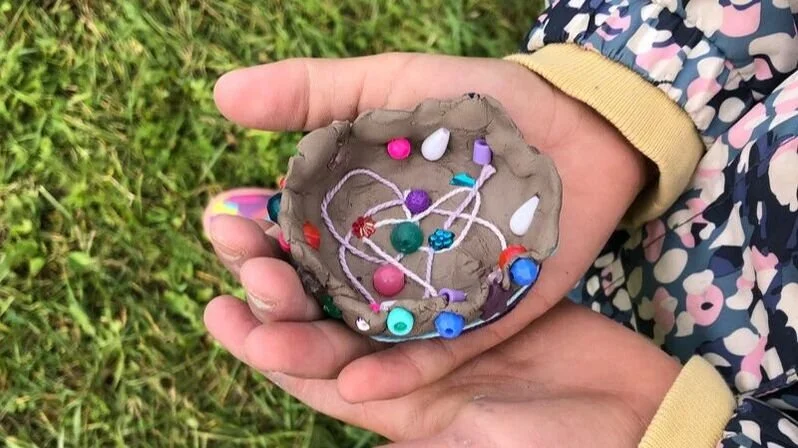 A child's hand holding a small, handmade clay dish with various jewelry pieces, including beads, charms, and thread, outdoors on grass.