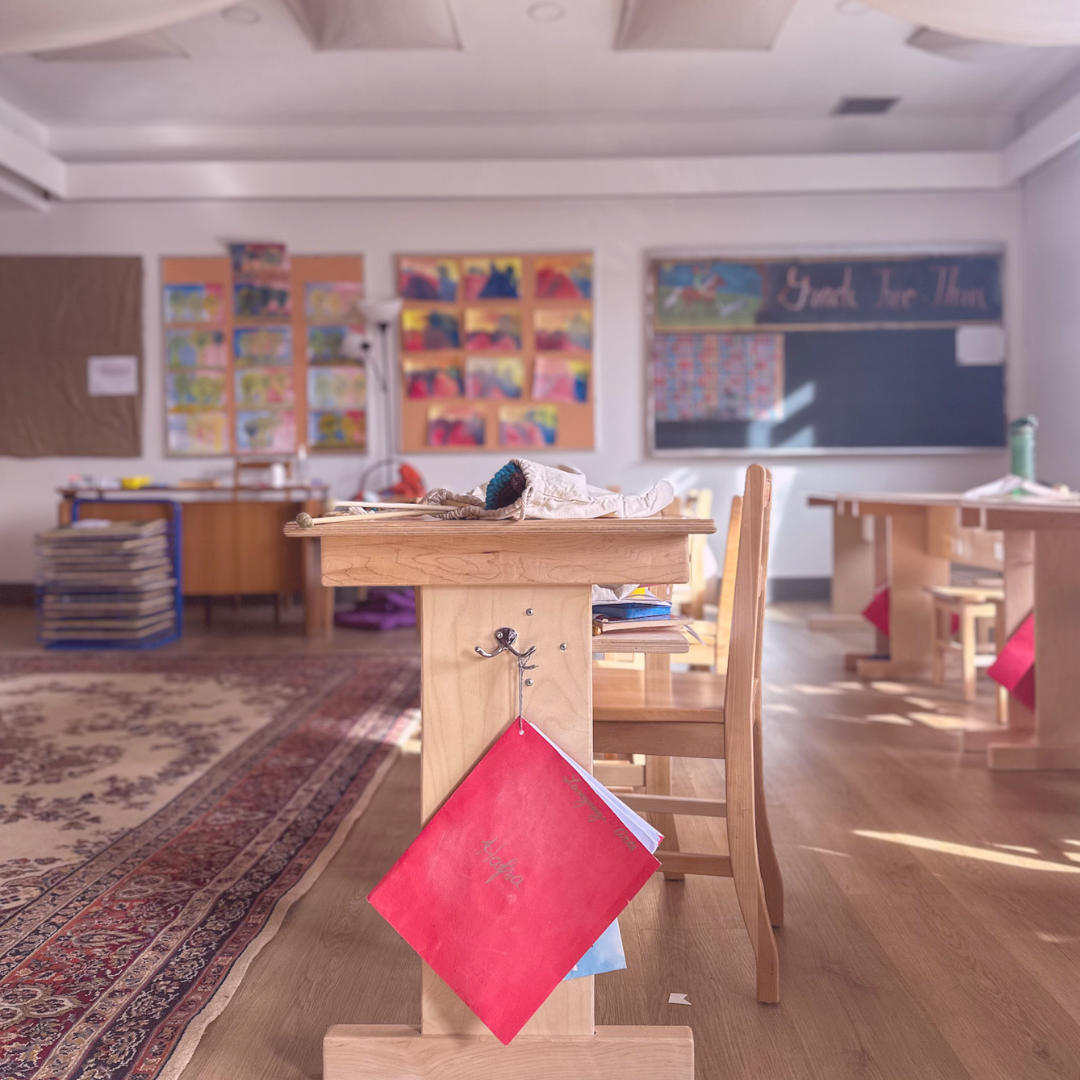 A classroom with wooden desks and chairs, colorful artwork on the walls, and a blackboard that says 'Thank You Than' in the background. There are supplies and a water bottle on the desks, with sunlight streaming in.