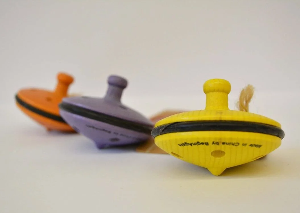 Three colorful spinning top toys in yellow, purple, and orange, lined up on a white surface with a plain background.