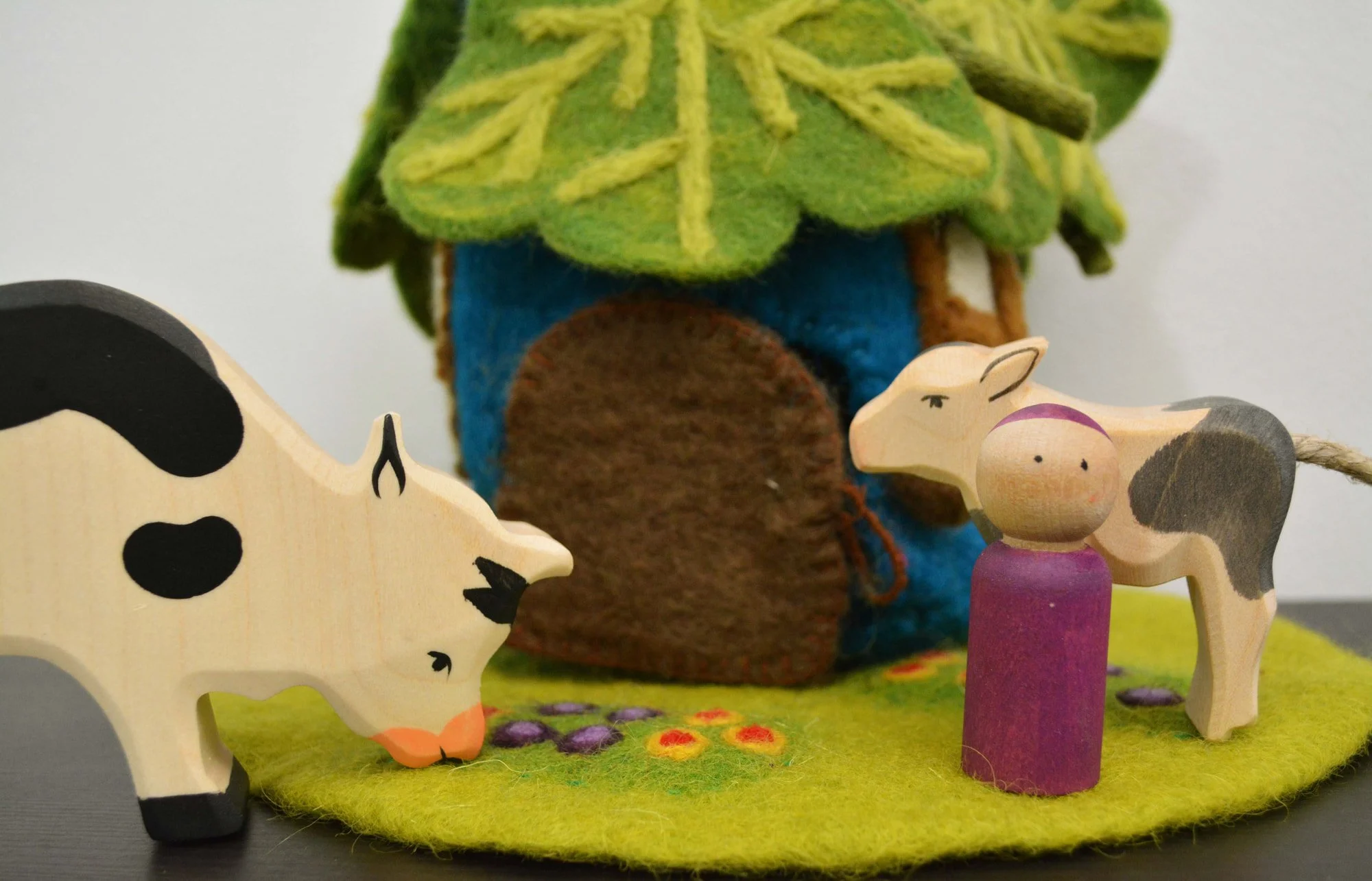 Felted toy forest scene including a felt tree, a wooden pig figure, a white and black spotted cow, a wooden pig figure, and a purple felted fence on a green felt base.
