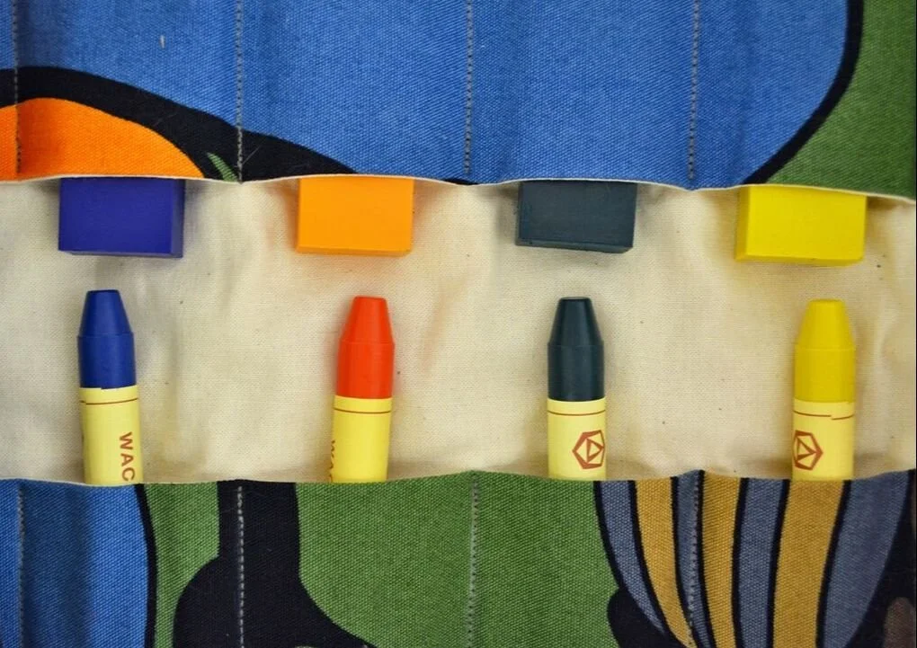 Four yellow markers with blue, orange, black, and yellow caps, each with colored ink, arranged in a line on a fabric case with colorful patches.